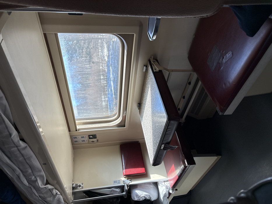 The image depicts the interior of a train compartment, likely traversing through or near the city of Vyazovaya, Russia. The scene is devoid of people.
In the upper left, a bunk bed is visible, covered with a folded white and grey striped blanket. Below it, an off-white wall panel features a small, red upholstered backrest and a corresponding red upholstered bench seat with a light beige base. A black bag and a white, plastic-wrapped object rest on the leftmost part of this seat. Numbered placards "52" and "51" are affixed to a metal support structure beside the seat.
Centrally, a rectangular table with a grey speckled surface and dark brown trim is extended, supported by a folding mechanism. Sunlight reflects off its surface. Below the table, a beige heating element with a gridded vent is visible.
To the right of the table, a window framed in light beige reveals a winter landscape outside. The exterior view shows snow-covered ground and a dense forest of bare trees under a bright, possibly overcast, sky.
Below the window and to the right of the table, another red upholstered bench seat with a light beige base is present. A light-colored smudge or mark is visible on its surface. Above this seat, a small, grey metallic cup holder is mounted on the wall. The compartment floor is dark grey. The overall lighting is bright, primarily from the sunlight entering through the window.
