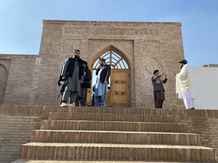 The image displays an outdoor scene at the entrance to the Ahmad Shah Durrani Shrine in Kandahar, Afghanistan, under a clear blue sky. A set of light brown brick stairs, comprising at least five visible steps, leads up to a substantial brick structure. The building features an ornate arched entryway with carved wooden double doors and an arched window above them. Decorative geometric patterns are integrated into the brickwork of the facade.
Five adult males are positioned on the upper stair landing and adjacent platform. On the left side of the platform, a bearded man with glasses, dressed in a dark grey shalwar kameez and a dark sleeveless vest, stands holding a black assault rifle slung across his chest, pointed downwards. Two other men are partially visible behind him and to his right; one wears a light blue shalwar kameez and a dark jacket. On the right side of the platform, a bearded man in a brown shalwar kameez and dark vest holds a black smartphone horizontally, pointed towards another heavily bearded man. This second man wears a white shalwar kameez and a white shawl draped over his shoulders and is gesturing with both hands, seemingly interacting with the smartphone user. The scene is brightly illuminated by direct sunlight.