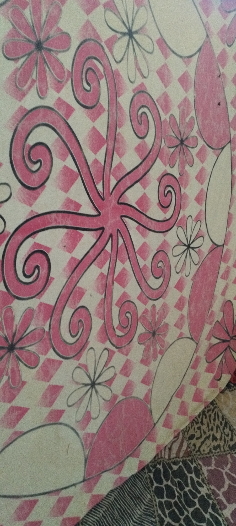 The image displays a close-up view of a circular object, possibly a table or a decorative piece, adorned with a pink and white diamond pattern background. Overlaid on this pattern are stylized pink floral elements and black line drawings of swirls and petals. The object appears to be made of a smooth material, possibly painted wood or laminate. The lighting is diffuse, and there is a slight blur to the image, suggesting it was taken handheld. The bottom right corner of the image shows a textured surface with a patchwork of animal prints, including leopard, zebra, and giraffe patterns. This suggests the object is placed on or near a surface with this distinct pattern. There are no people or specific landmarks visible that would indicate a location in Yaroslavskaya, Russia.