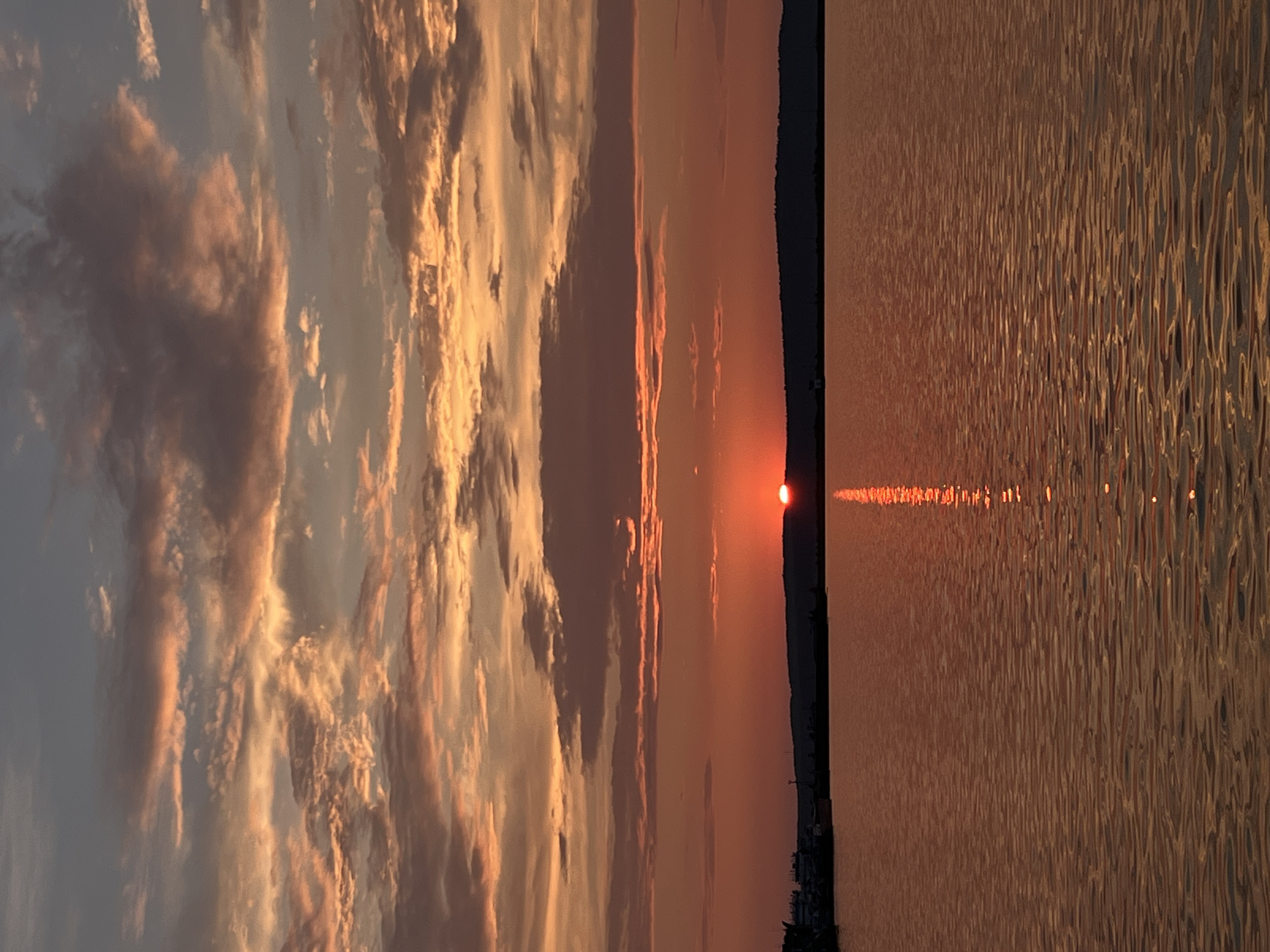 A serene sunset over the Kama River in Chaikovsky, Russia, paints the sky with hues of orange and pink, reflected on the calm water's surface. The sun, a vibrant orb, is partially obscured by a distant, silhouetted treeline, casting a shimmering path of light across the river. On the left bank, a cluster of industrial buildings and docked vessels suggests the presence of a port or shipyard. The sky is adorned with scattered clouds, catching the last rays of sunlight, adding depth and texture to the twilight scene. The overall impression is one of tranquility and natural beauty, with a hint of industrial presence at the edge of the landscape.