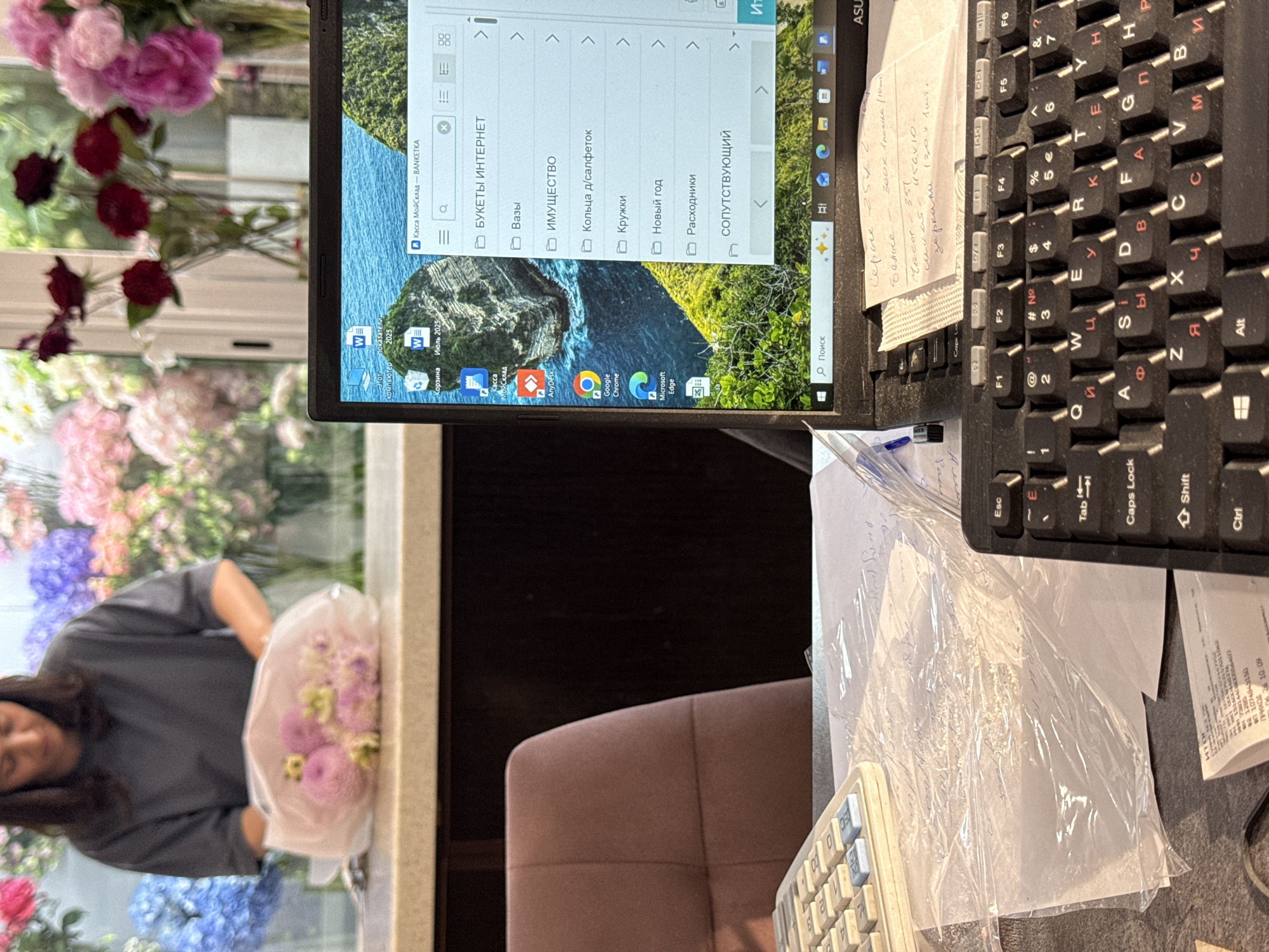 This is a close-up, high-angle shot of a woman arranging a bouquet of flowers. She is wearing a grey t-shirt and has dark hair. The flowers are light pink, purple, and white, and are wrapped in clear plastic. In the background, there's a laptop displaying a file explorer window with Russian text, possibly indicating a business or organization named "Касса МойСклад" (Cashier MyWarehouse). The laptop screen also shows a desktop wallpaper of a mountainous landscape. To the right of the laptop is a keyboard with Russian lettering. A handwritten note is attached to the keyboard, with some prices and item descriptions visible in Russian. The overall scene appears to be indoors, possibly a shop or a home office, with a focus on floral arrangements and work-related tasks. The lighting suggests it could be daytime.