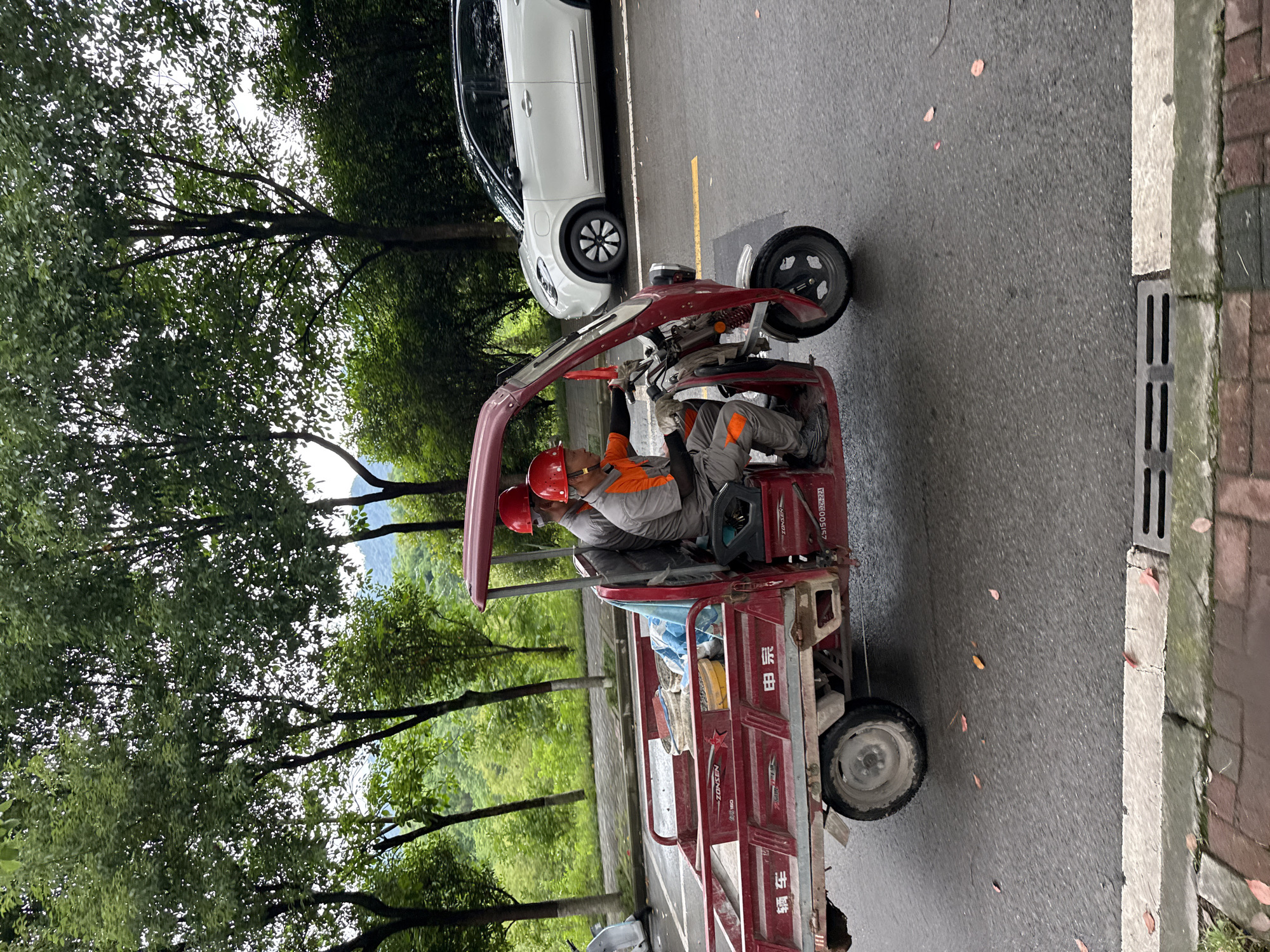 Two men in orange and grey uniforms and red helmets are riding in a red electric tricycle on an asphalt road. The tricycle has a small front cab and a larger open cargo area with blue and white items in the back. The characters "电宗” are on the side. A white car is parked on the side of the road, separated by a yellow line. Trees and a green hillside are on the left. A brick and concrete curb with a metal drain is on the right. The location is likely Jiangjin, China based on the script on the tricycle.