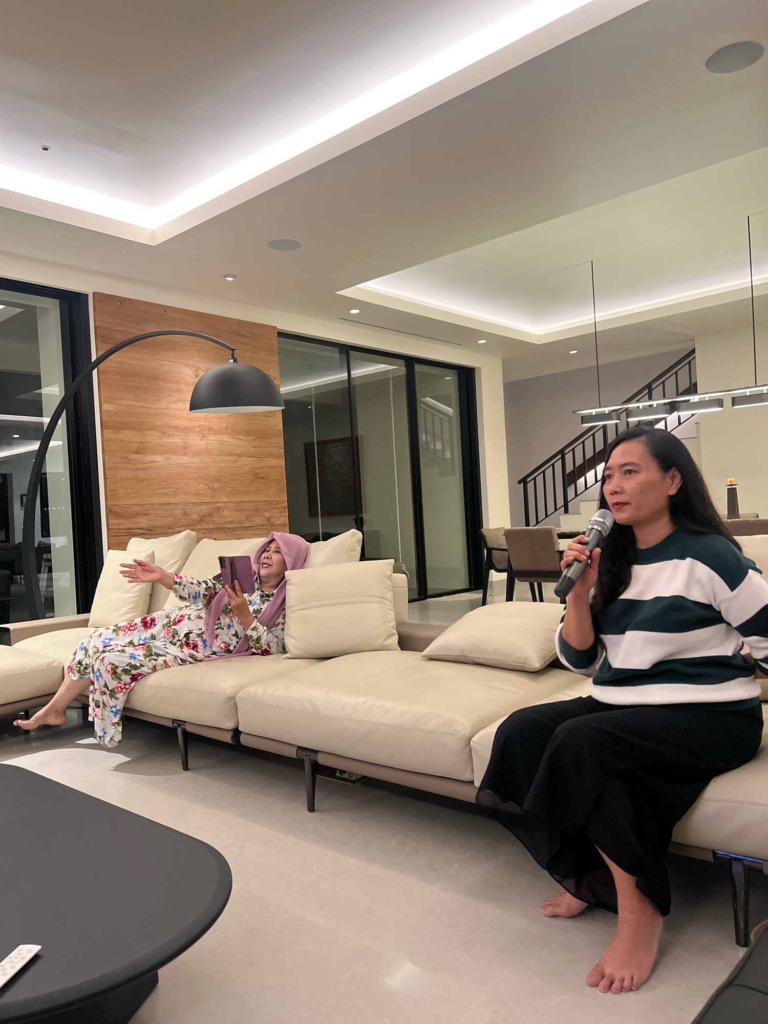 Two women are in a modern living room in Ngaglik, Indonesia. One woman, wearing a floral-patterned pajama set and a light purple hijab, is sitting on a light-colored sectional sofa and looking at a tablet. The other woman, wearing a green and white striped sweater and black pants, is sitting on the same sofa and holding a microphone. She appears to be singing or speaking into the microphone. The living room has a wooden accent wall behind the sofa, a large arc lamp, and a ceiling with recessed lighting. A dark gray coffee table with a remote control is in the foreground. The background shows a staircase and dining area.