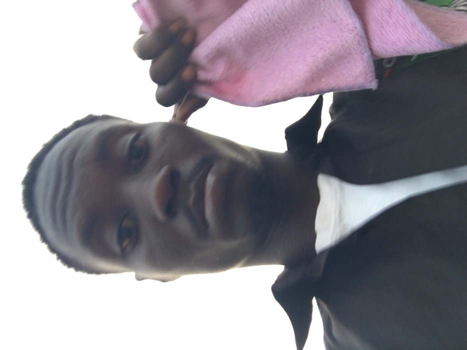 The image captures a close-up portrait of a dark-skinned person, likely male, in an upward-angled shot. The individual's head is tilted, and they gaze directly at the camera with a neutral expression. Visible from the upper chest, the person wears a dark-colored collared shirt, possibly black or dark brown, layered over a white undershirt or collar.

A hand, belonging to the subject, is positioned near their head, holding a piece of soft, pink fabric, which appears to be a towel or cloth. The fabric is gathered in the fingers. The background is entirely bright white, suggesting either a heavily overexposed sky or a plain white backdrop, consistent with an outdoor setting under bright daylight conditions. There are no specific location cues visible within the frame, but the image is contextually located in Pindiga, Nigeria. No visible text is present in the image. The overall impression is that of a casual portrait or selfie.