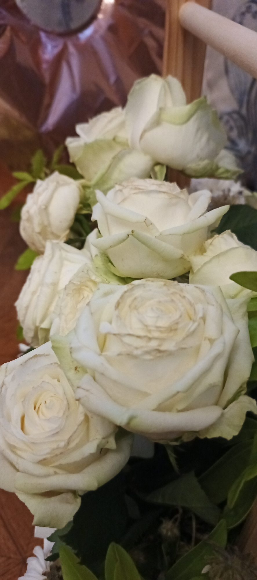 A bouquet of white roses is displayed in a vertical, close-up shot. The roses are in various stages of bloom, with some fully open and others still in bud. The petals are delicate and show slight browning on the edges. Green leaves are interspersed among the flowers. The background is blurred, suggesting an indoor setting with a wooden surface and possibly fabric. There is no discernible indication of the city of Samara, Russia.