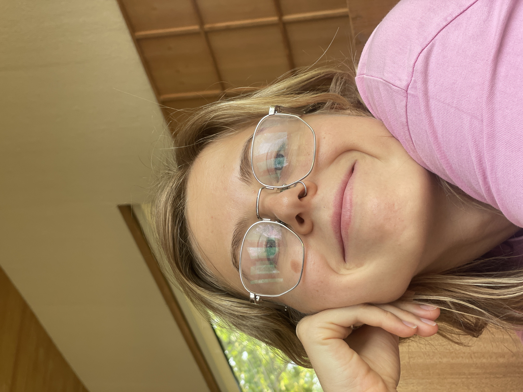 A person with blonde hair and blue eyes, wearing silver-framed glasses and a pink shirt, is resting their head on their hand. The background includes a wooden ceiling, a tan wall, and greenery visible through a window. There is no information indicating the location is in Chiang Mai City Municipality, Thailand.