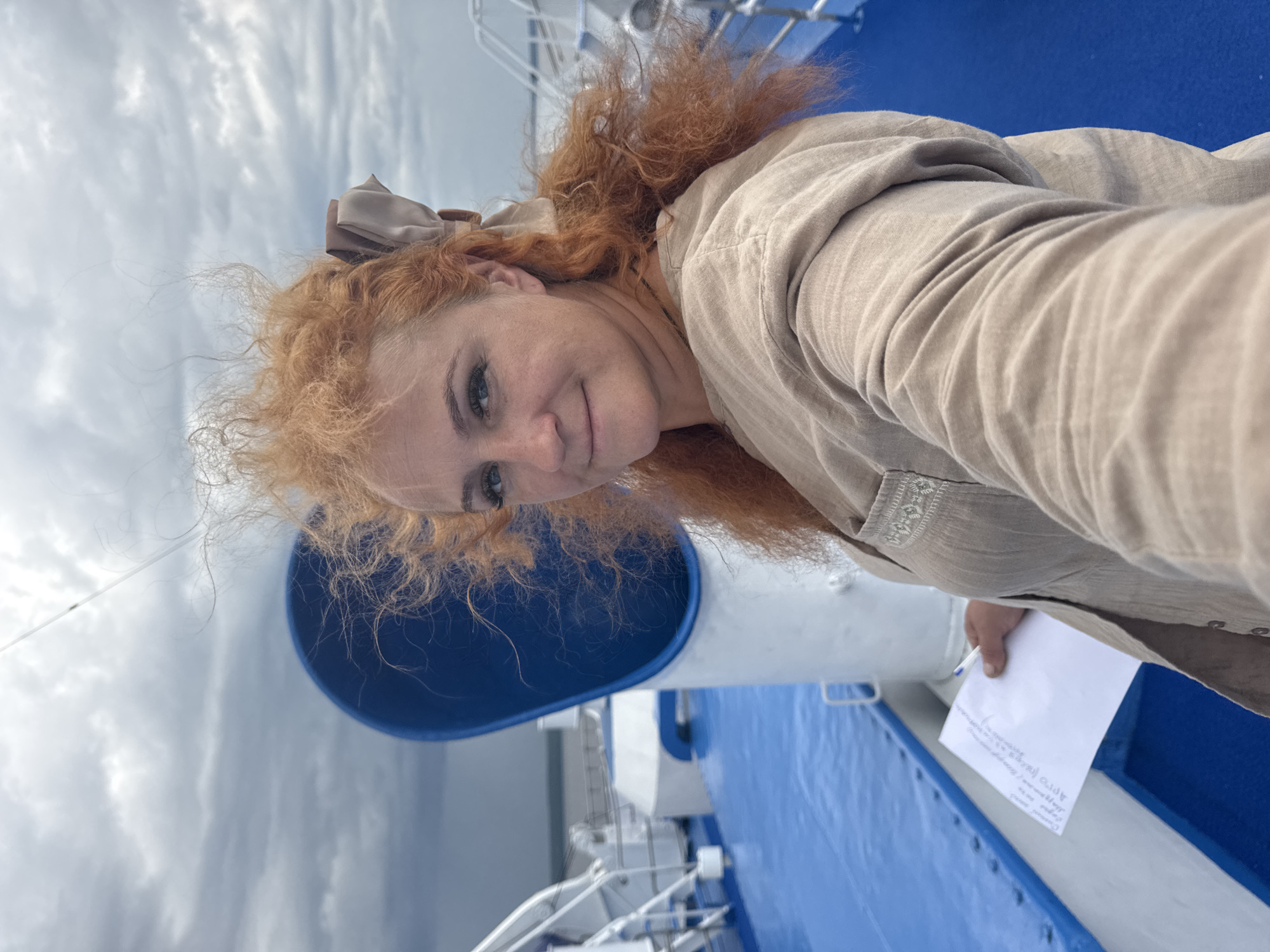 A medium-shot, selfie-style photograph captures a woman with vibrant red, curly hair tied back with a beige bow, standing on the deck of a vessel. She is looking towards the camera with a slight smile and her head is tilted. The background shows the blue deck of the ship, white railings and structures, and a cloudy sky. A large blue, circular object is visible behind her head. In her right hand, she holds a white piece of paper with some handwritten text and a pen. The woman is wearing a light brown, linen-like garment with decorative stitching on the chest area. The environment suggests an outdoor setting on a boat, possibly on a body of water. The location is identified as Ikskoe Ust'e, Russia, which is a port located at the confluence of the Lena River and the Ust-Kutka River.