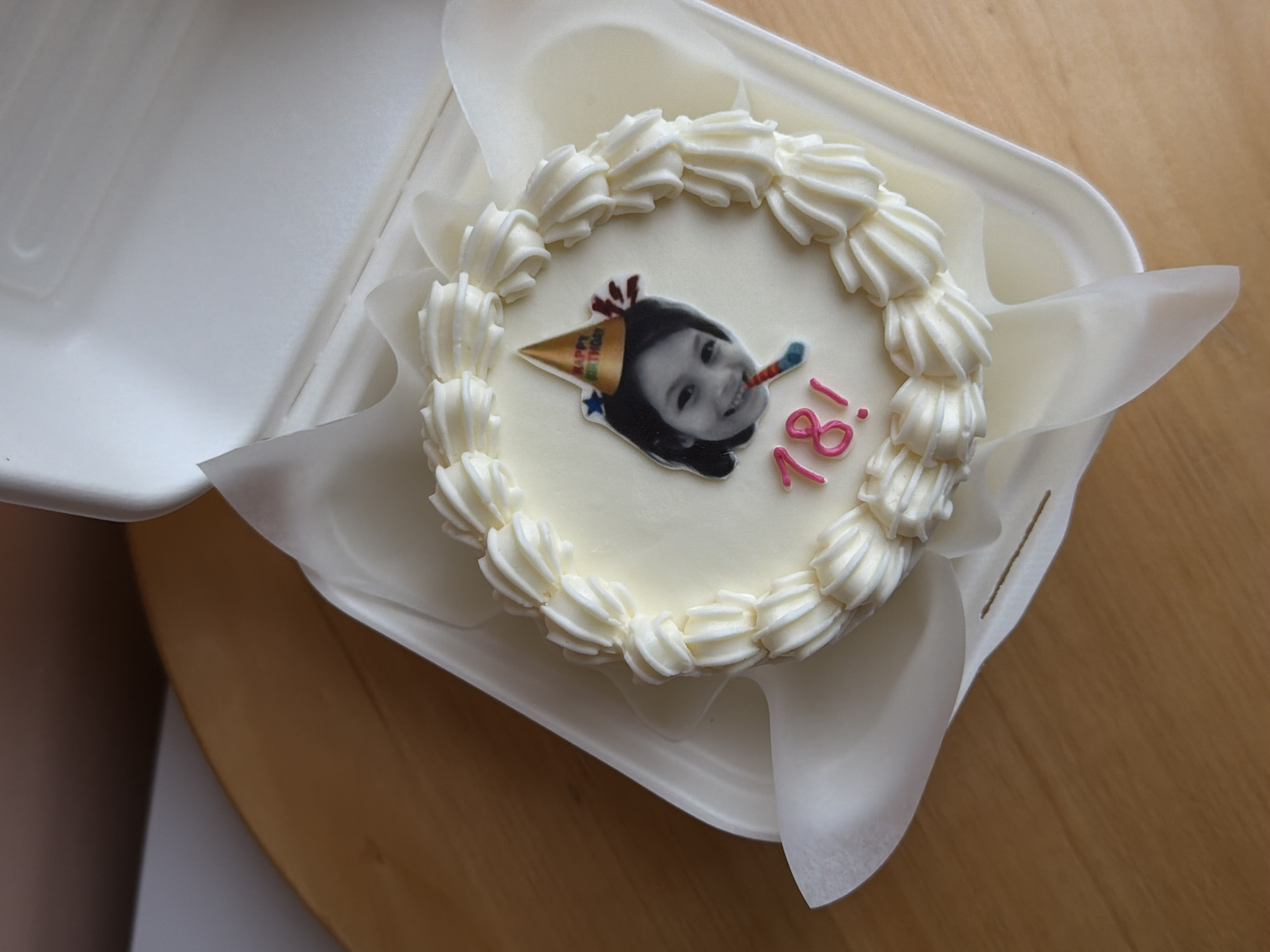 The image shows a round cake in a white rectangular container. The cake is decorated with white frosting around the edge and a small picture of a person wearing a party hat and blowing a noisemaker. "18!" is written on the cake in pink. The container is lined with white paper. The cake is sitting on a wooden surface. There is no evidence to suggest the location is Tel-Aviv, Israel.