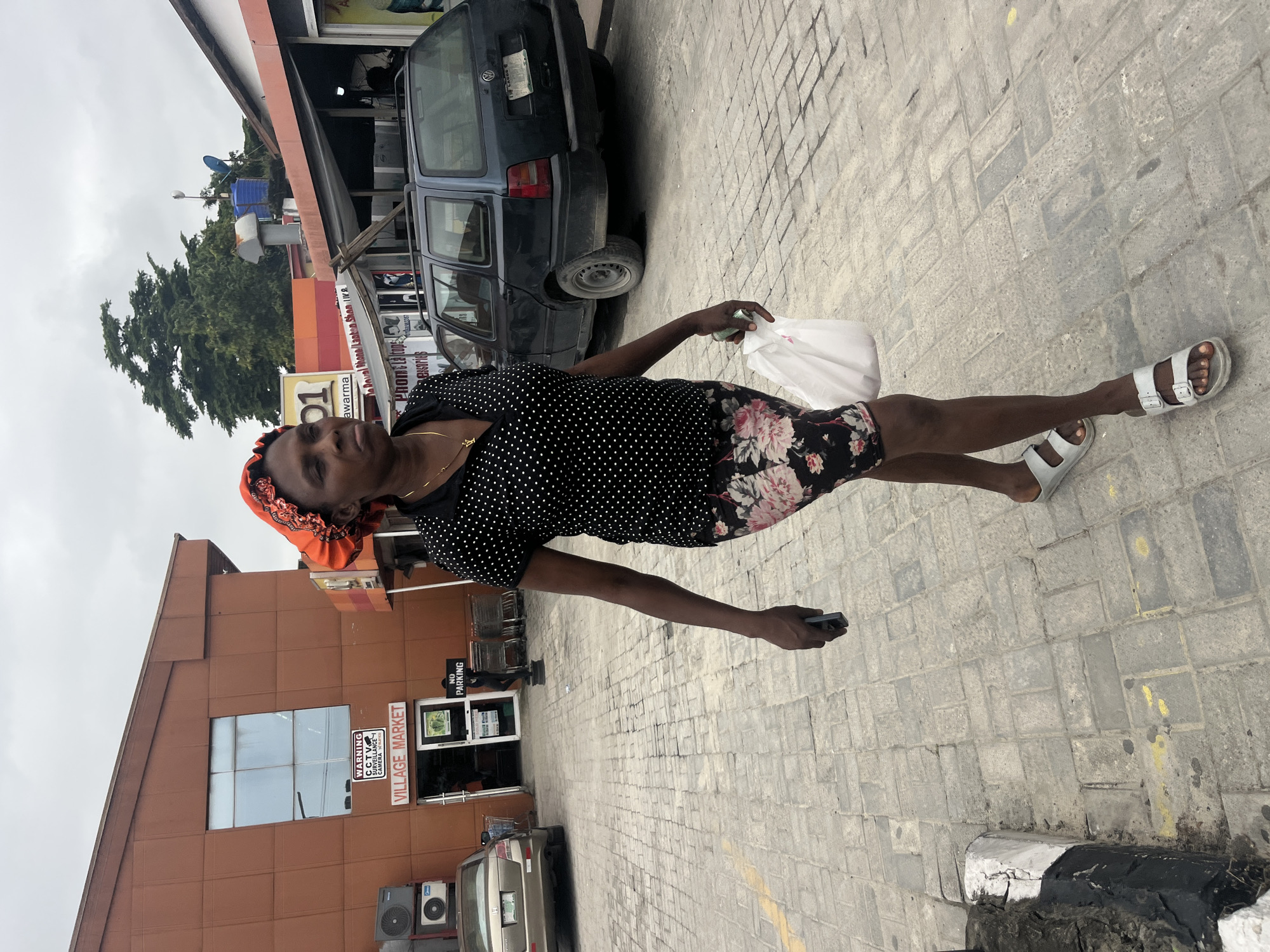 In Ado, Nigeria, a Black woman walks on a paved sidewalk in front of a building labeled "VILLAGE MARKET." She wears a red headscarf, a black polka-dot shirt, and a floral-patterned skirt. She carries a white plastic bag in her right hand and a smartphone in her left. Behind her, a dark blue station wagon is parked. The sky is overcast.
