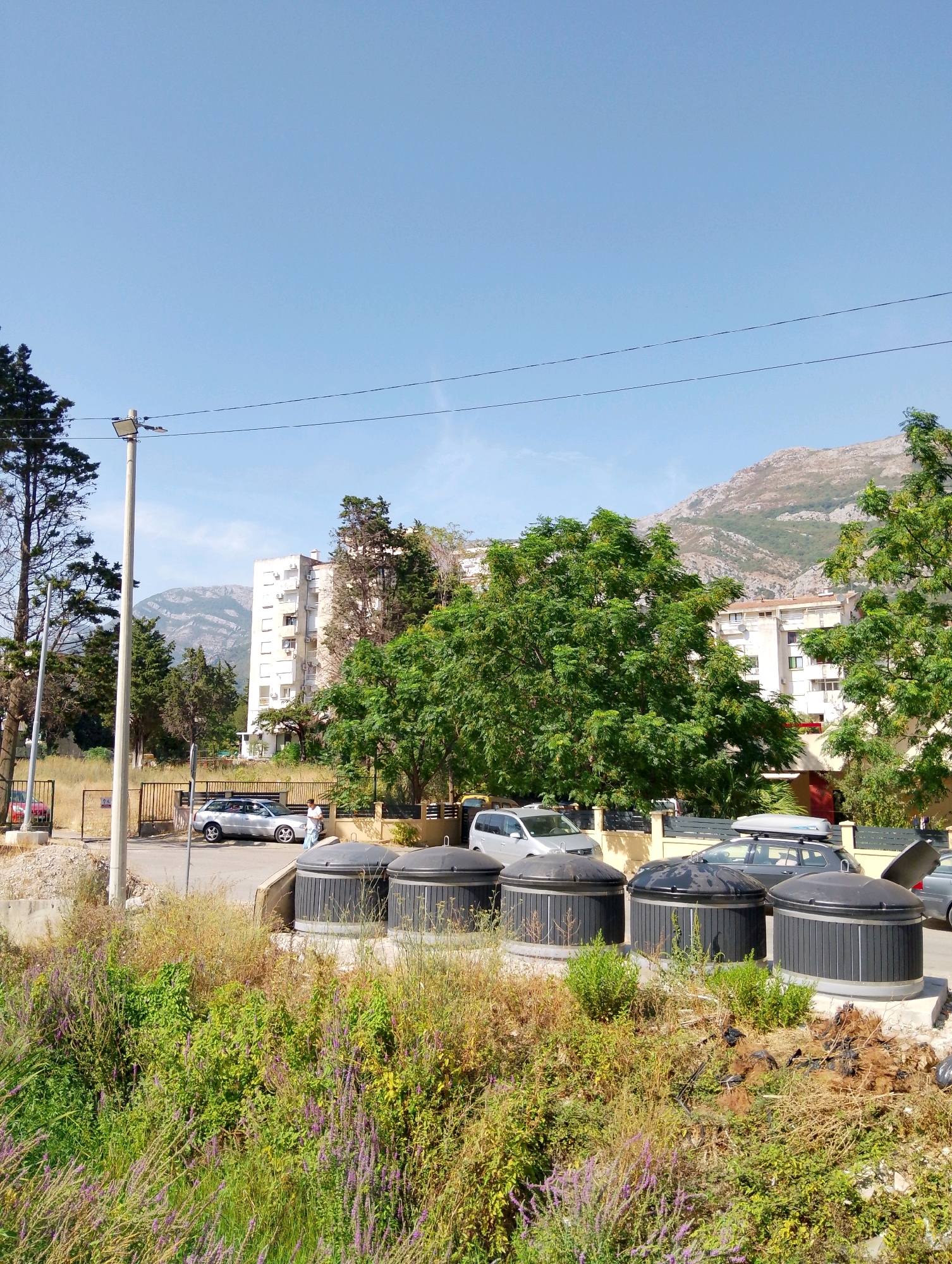 In the city of Sutomore, Montenegro, a bright, clear sky hangs over a scene depicting an outdoor area with overgrown vegetation in the foreground. Several large, dark-colored cylindrical trash receptacles with black lids are lined up on a concrete base. Behind the receptacles, a low fence separates the area from a paved street. On the street, a silver station wagon is parked, and a person in a white shirt and blue pants stands near it. Further back, a multi-story apartment building with numerous windows and balconies stands prominently, surrounded by lush green trees. In the distance, a majestic mountain range rises against the horizon. A lamppost with a light fixture is visible to the left of the frame, and power lines stretch across the sky. The overall impression is a blend of urban infrastructure and untamed natural elements.