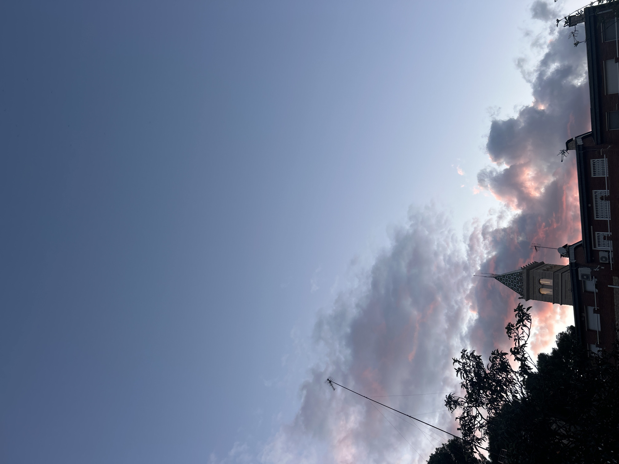 The photograph captures a dusk or dawn scene in Barcelona, Spain. A building is visible on the right, partially obscured by silhouetted trees and a network of antennae. The building features a tower-like structure with ornate detailing. The sky transitions from a clear blue on the left to a band of clouds tinged with pinks and purples on the right, likely illuminated by the rising or setting sun.
