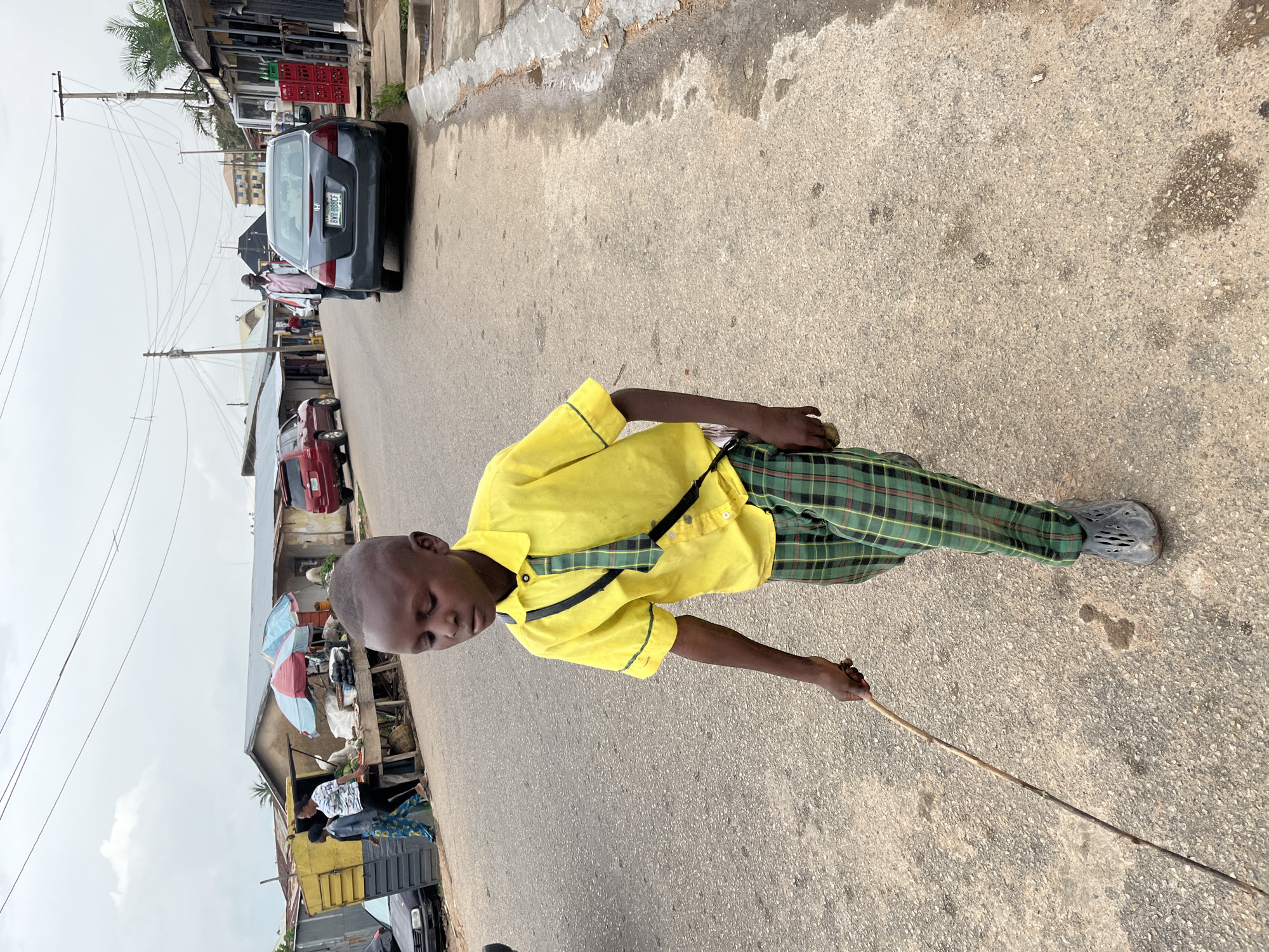 The image depicts a vibrant street scene in Jos, Nigeria, during the daytime under an overcast but bright sky. The central subject is a young boy, possibly a student, with a shaved head, wearing a bright yellow short-sleeved shirt with green trim, green and black plaid trousers, and dark grey slip-on shoes. A black strap is visible across his shoulder. He walks along the edge of an asphalt road, holding a long, thin stick in his right hand, pointing it towards the ground. His head is bowed, looking downwards.
In the background, the street is lined with various structures, including simple buildings and market stalls. Two distinct umbrellas, one blue and one pink/red, are visible over what appears to be market activity. Several people are scattered in the background, some near these stalls. A dark grey sedan, possibly a Honda Accord, is parked on the side of the road with a visible license plate: "RV 880 GCC." Other vehicles, including a red car and a dark truck, are partially seen further down the street. Overhead, numerous utility poles with crisscrossing wires are prominent against the pale sky, contributing to the urban landscape. The scene conveys a typical, bustling street environment.