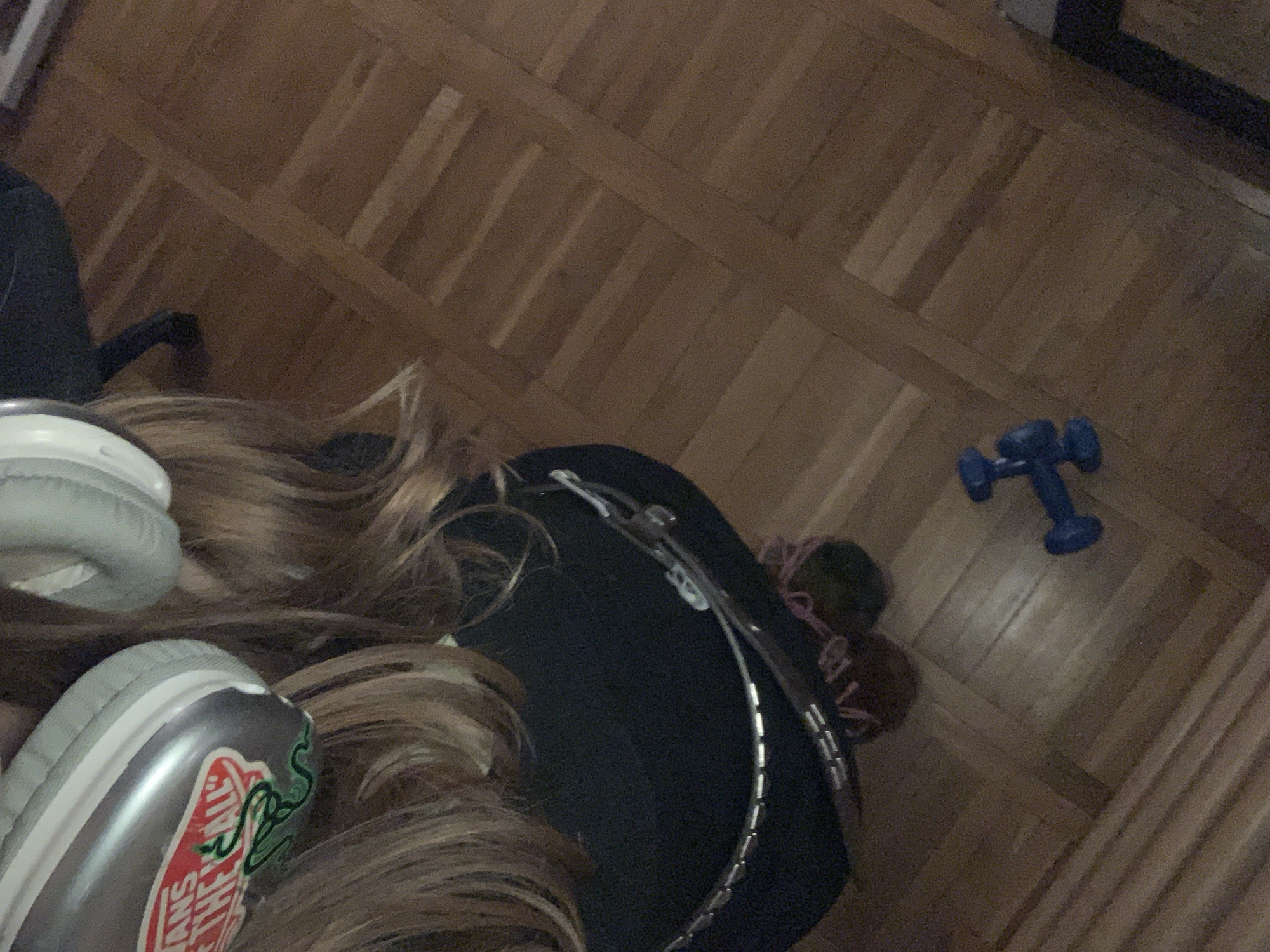 A top-down shot shows a section of a wooden parquet floor in a home setting. A person with long, light brown hair is partially visible, with headphones and a silver Vans shoe with a logo partially obscuring the hair. A black item (possibly clothing or a bag) is present, adorned with a metallic chain-like accessory. To the right of the person, on the floor, is a blue dumbbell. It is not possible to determine a specific location within Vozdovac Urban Municipality from the given image.