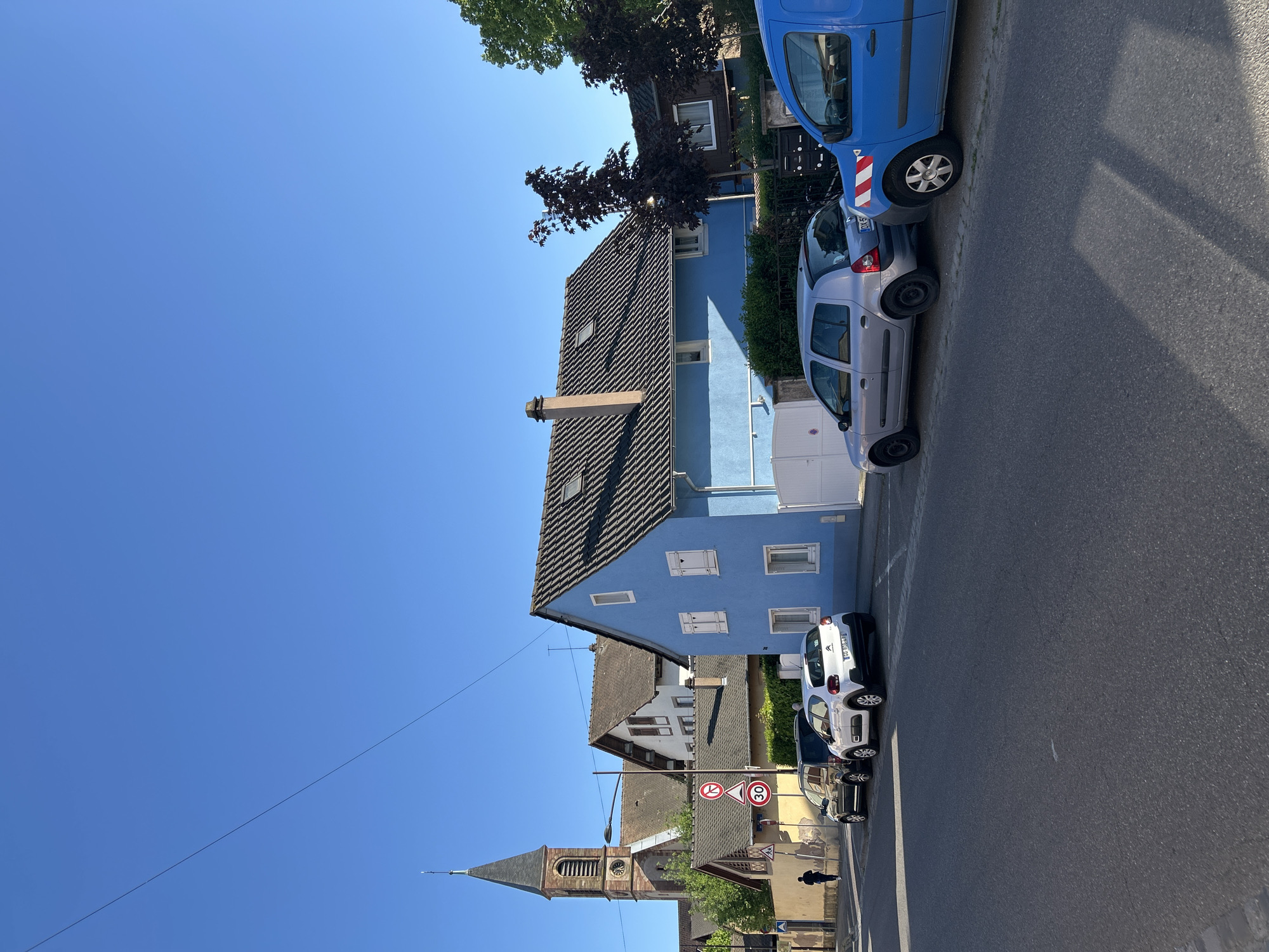 This image captures a vibrant, sunlit street scene in Strasbourg, France, under a clear, bright blue sky. The focal point is a distinctive two-story house painted a light, vivid blue with white window frames and shutters, and a dark tiled roof featuring a chimney. Several other residential buildings with varied architectural styles, including one with brown shutters, are visible further down the street.
Along the right side of the asphalt road, multiple cars are parked. These include a blue commercial van with white and red striped markings, a silver hatchback car (its license plate partially visible, starting "DA-5..."), a white hatchback (possibly a Citroën C3), and a darker-colored car. Bright sunlight casts distinct shadows from the parked vehicles and overhead trees onto the road, indicating it is daytime, likely mid-morning or early afternoon, with excellent weather conditions.
In the distance, to the left, a traditional church with a tall, pointed steeple, a clock face, and arched windows adds a historical element to the urban landscape. Green and dark-leafed trees are scattered throughout the scene. Road signs include a circular sign indicating a "30" km/h speed limit, a "No Entry" sign (red circle with white bar), and a triangular "Warning" sign. A solitary figure appears to be walking on the sidewalk in the lower-left section of the image. The scene portrays a typical, peaceful day in a European town.