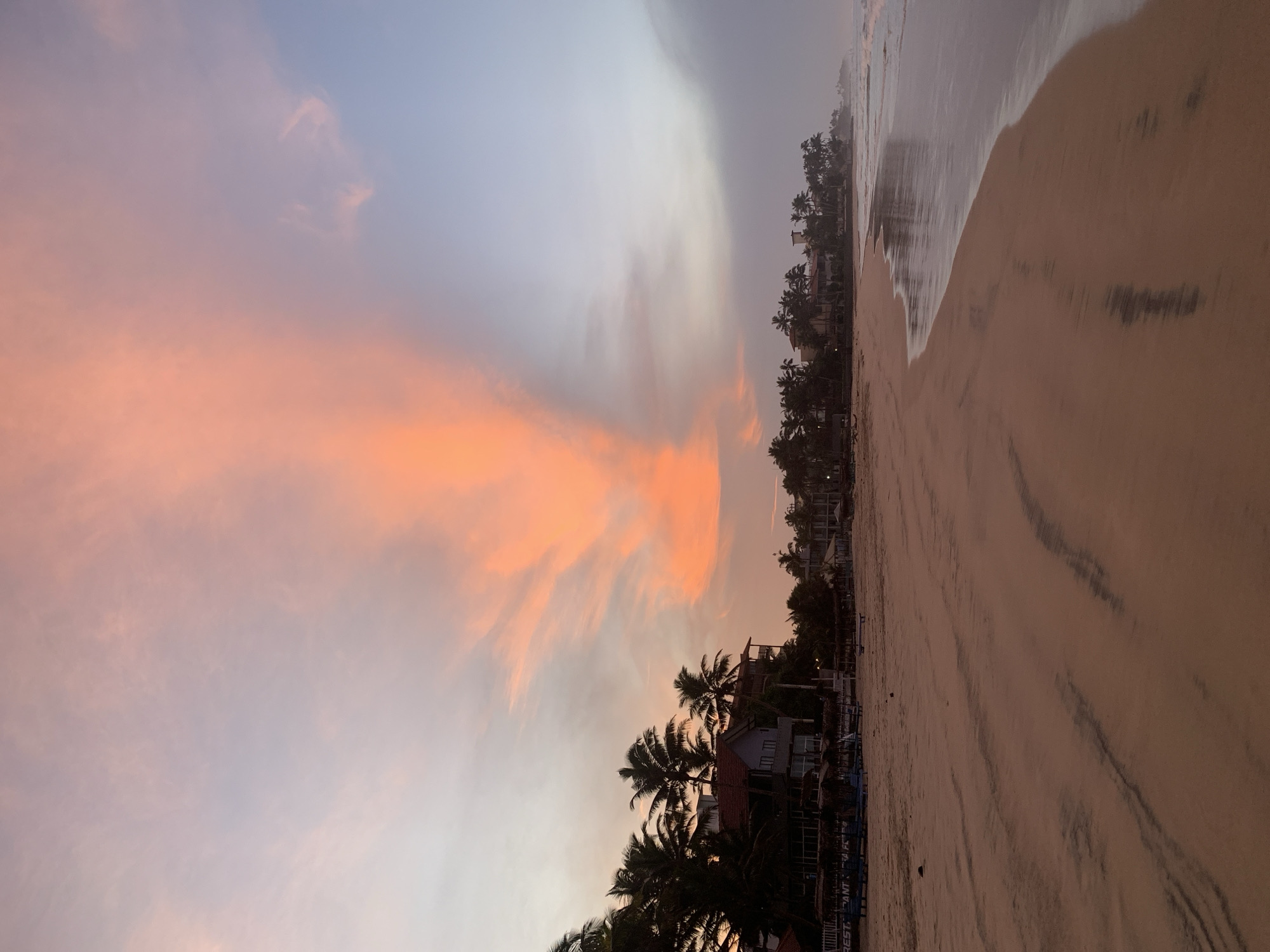 The image depicts a beach at dusk in Kathaluwa, Sri Lanka. The sky is filled with orange and grey clouds. The beach extends from the lower right to the upper right and is lined with palm trees and low-rise buildings. The tide is coming in, with the ocean surface reflecting the colors of the sky.