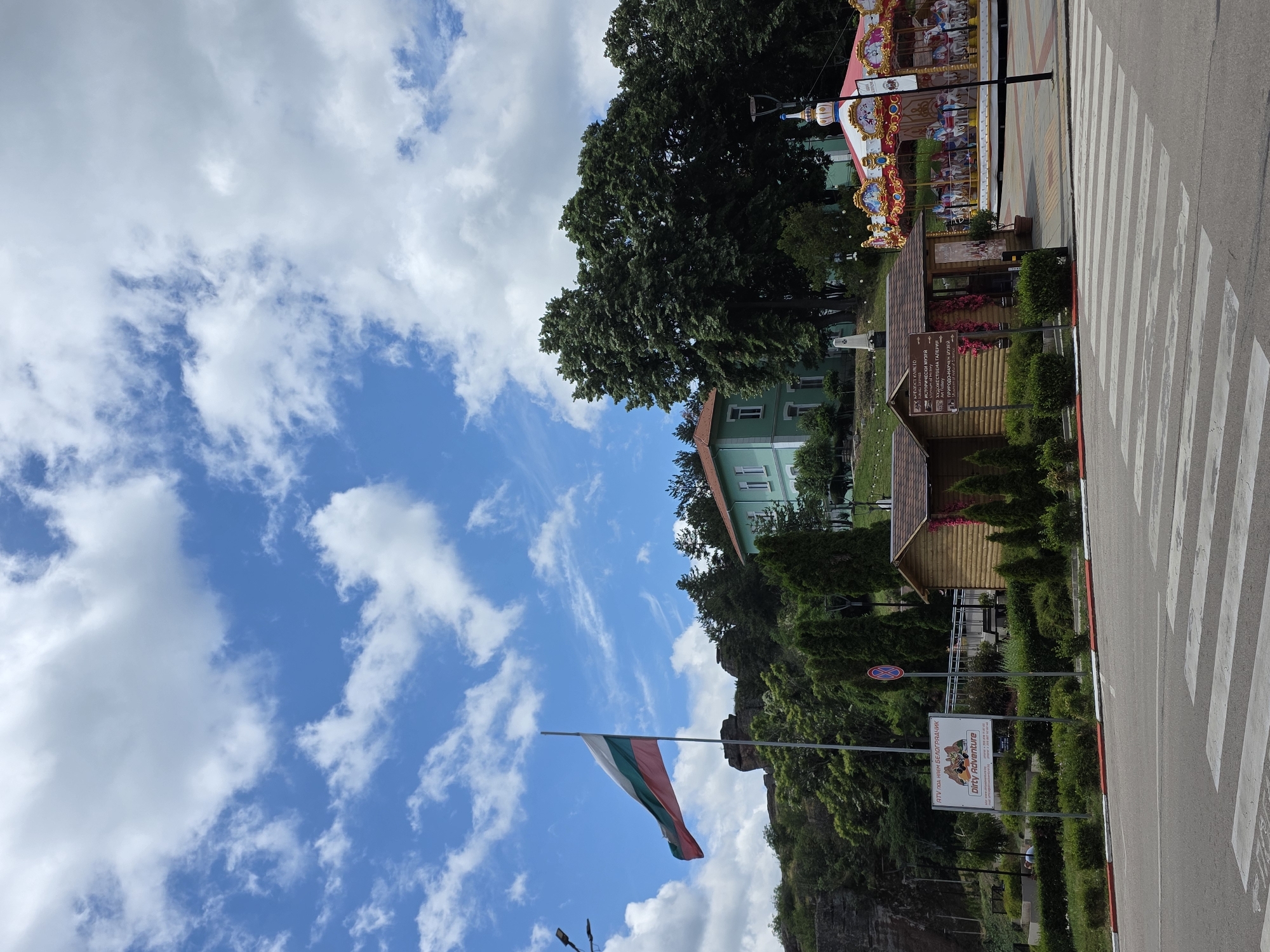 The image captures a scene in the city of Belogradchik, Bulgaria, featuring a large Bulgarian flag waving in the foreground against a backdrop of a bright blue sky with scattered white clouds. To the left of the flag is a rocky cliff formation, and further to the right is a light green building with a red-tiled roof, likely a public building. In the mid-ground, there are several small wooden structures, one of which appears to be a kiosk or information booth, and a colorful carousel. The scene also includes trees, bushes, and a street with a pedestrian crossing in the lower part of the image. A sign with Bulgarian text and the words "DIRTY ADVENTURE" is visible on the left side. The overall impression is a public space, possibly a park or town square, during daytime.