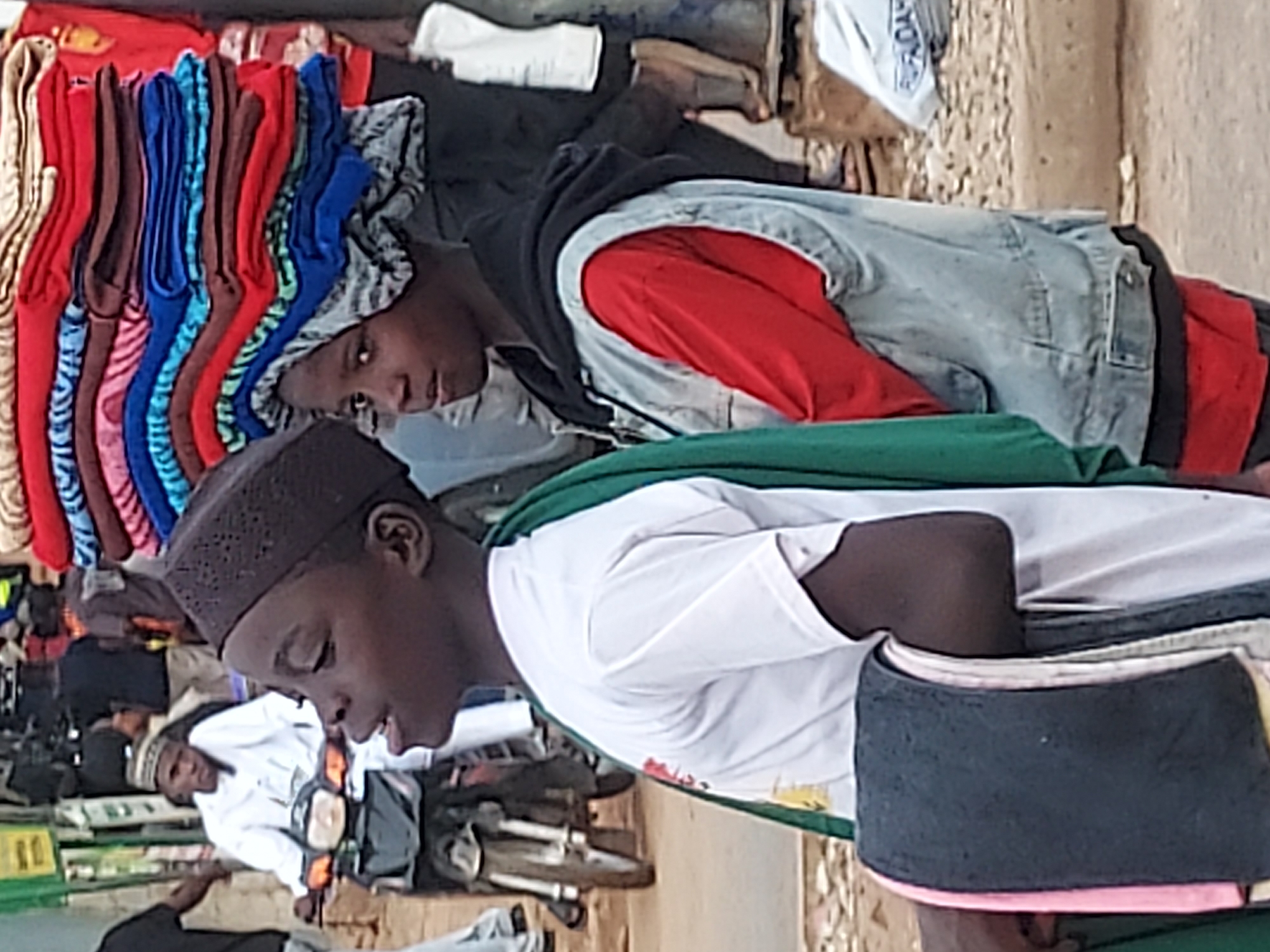 This image captures a vibrant street or market scene in Jos, Nigeria, during daylight hours. The photo is taken from a slightly elevated perspective, looking down into the activity.
In the immediate foreground, on the left, a young person with dark skin is visible in profile, wearing a dark brown kufi (skullcap) and a white T-shirt with a green garment draped over their shoulders. They are looking downwards, seemingly focused on something out of frame. To their right, in the mid-ground, stands another young person with dark skin, wearing a black hoodie over a red long-sleeved shirt and a light blue denim vest. A light-colored, patterned head covering is visible under their hoodie. This individual is looking directly towards the camera with a neutral expression, positioned behind a neatly stacked display of colorful folded clothing, including items in red, brown, blue, pink, and patterned fabrics.
In the background, other people are visible, including one person seated on a motorcycle or scooter, wearing a white shirt and a light-colored hat. The setting features sandy or earthen ground, a light-colored wall or building in the distance, and various other market elements, suggesting a bustling commercial area. A white plastic bag in the background has some text, partially visible as "PRIDAY" or similar. In the bottom right corner of the foreground, part of a black and pink cap or hat is visible. The overall atmosphere appears active and typical of a busy outdoor market.