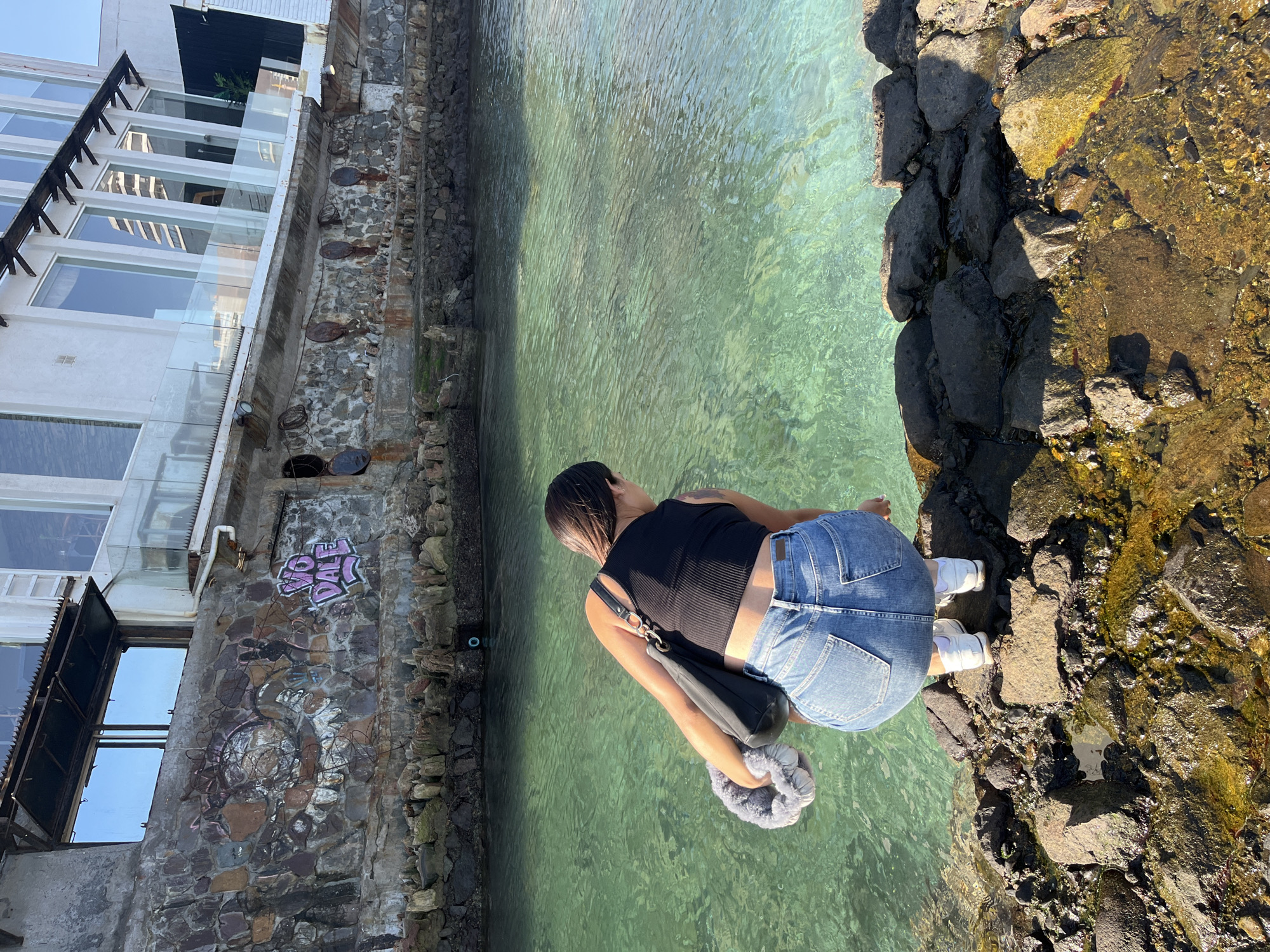 A young woman is bent over, looking into a pool of water. She is wearing a black tank top and blue jeans and has a purse slung over her shoulder. The water appears to be turquoise, and the surrounding area is rocky and covered with what looks like algae. To the left of the frame is a building with multiple windows and graffiti on the stone wall at the base. The location is likely a coastal area.