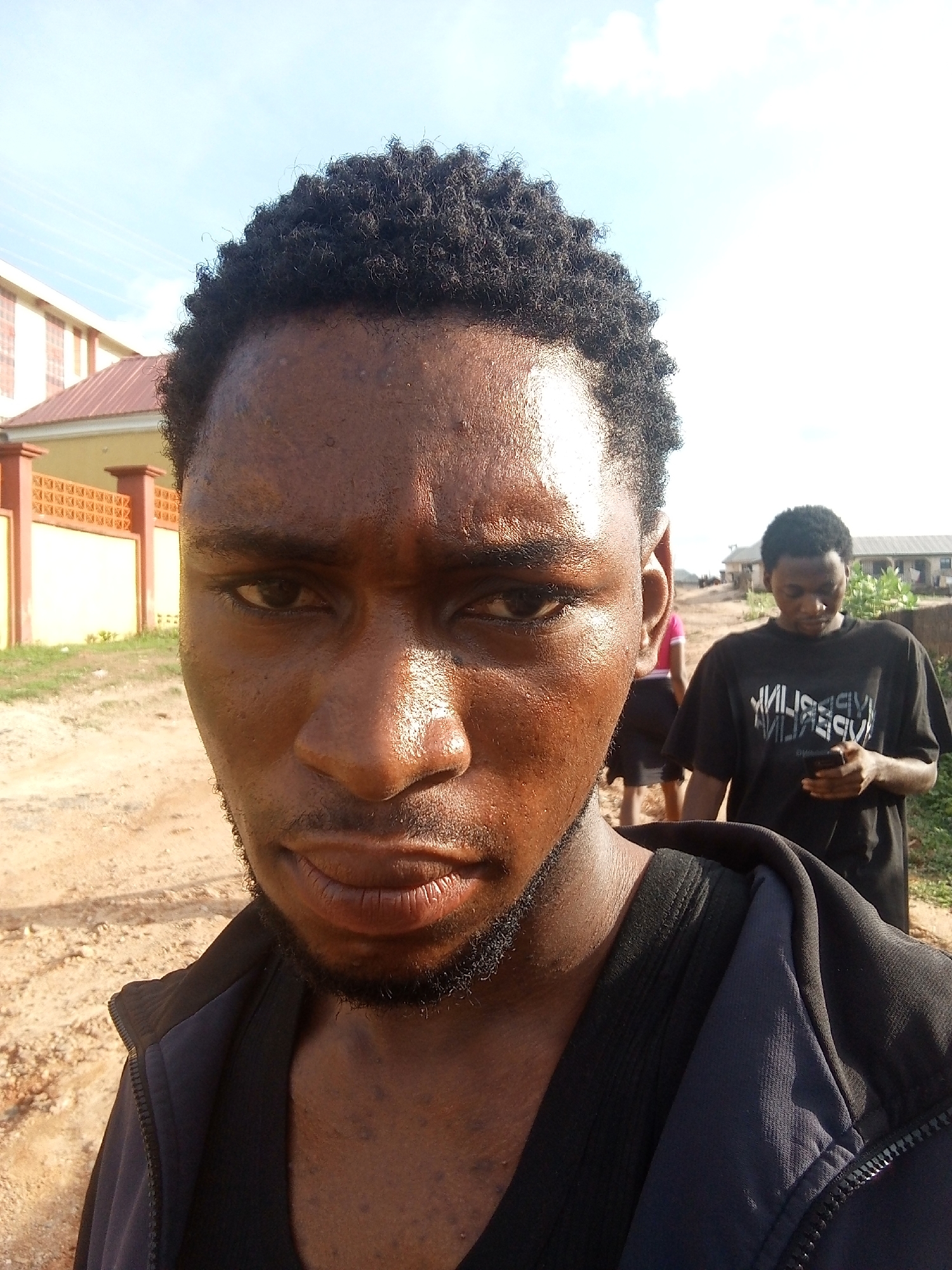 A close-up selfie shows a young, dark-skinned man with short, curly black hair and a short beard. His skin appears somewhat shiny, possibly due to sweat or oil. He is wearing a black t-shirt and a dark blue zip-up hoodie.
Behind him, a dirt road stretches into the background. Another young man, also dark-skinned, is standing on the road looking at a smartphone. In the background behind him, a building with a red roof is visible. To the left, there is a building with a yellow wall and a brick fence.
Based on the background and the presence of dirt roads, the location appears to be an area outside the main city center of Lokoja, Nigeria.