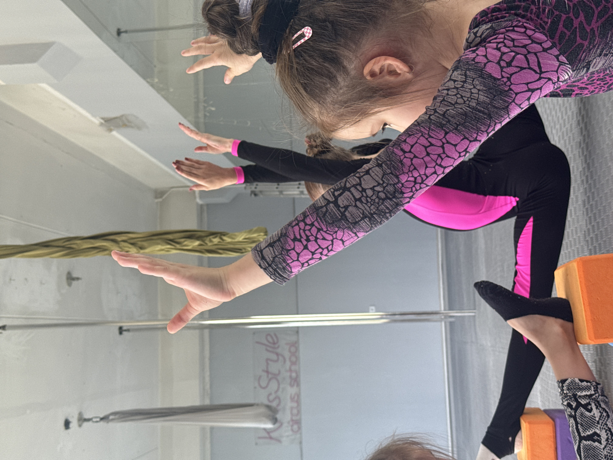 The image shows a group of young girls practicing aerial acrobatics in a studio. The girls are wearing leotards with a pink and black pattern and black leggings. One girl in the foreground is reaching out with her hands, and her legs are bent with her feet resting on orange blocks. Another girl behind her is in a similar pose. The studio has a reflective wall, showing a reflection of the ceiling and some aerial equipment hanging from above. The floor is light-colored, and there are metal bars and hanging silks visible. A logo for "KidsStyle Circus School" is visible on the wall, suggesting the location is a circus school. The overall impression is one of active learning and physical exertion in a dedicated training space.