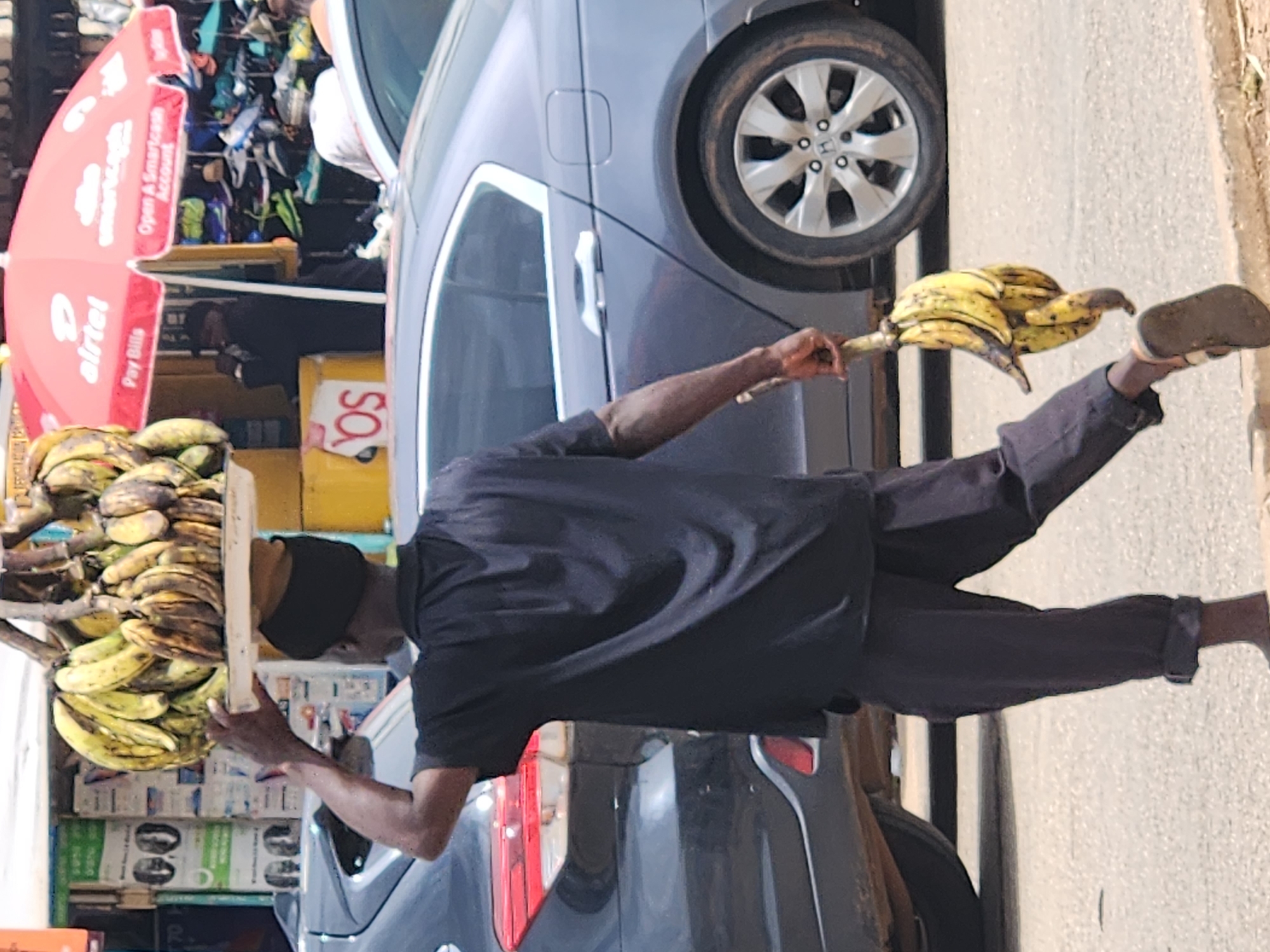 In Jos, Nigeria, a dark-skinned man walks with a bunch of plantains balanced on his head and another bunch in his right hand. He wears a black short-sleeved shirt and dark pants, with the cuffs rolled up. The man is barefoot on one foot and wearing a sandal on the other. He walks on a paved road, with a black SUV to his left and a gray sedan to his right. In the background, stalls with goods are visible, including one with a red umbrella advertising "airtel" and "smartcash." The scene appears to be a bustling marketplace.