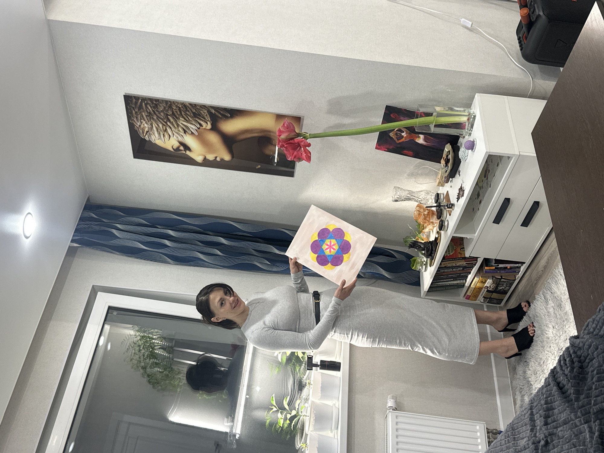 A woman in a grey dress and black shoes holds a square drawing with a flower design, standing indoors near a window with plants. Behind her is a white radiator, a cabinet with a black speaker on top, and wall decorations including a portrait and a flower in a vase. The location is Murino, Russia.