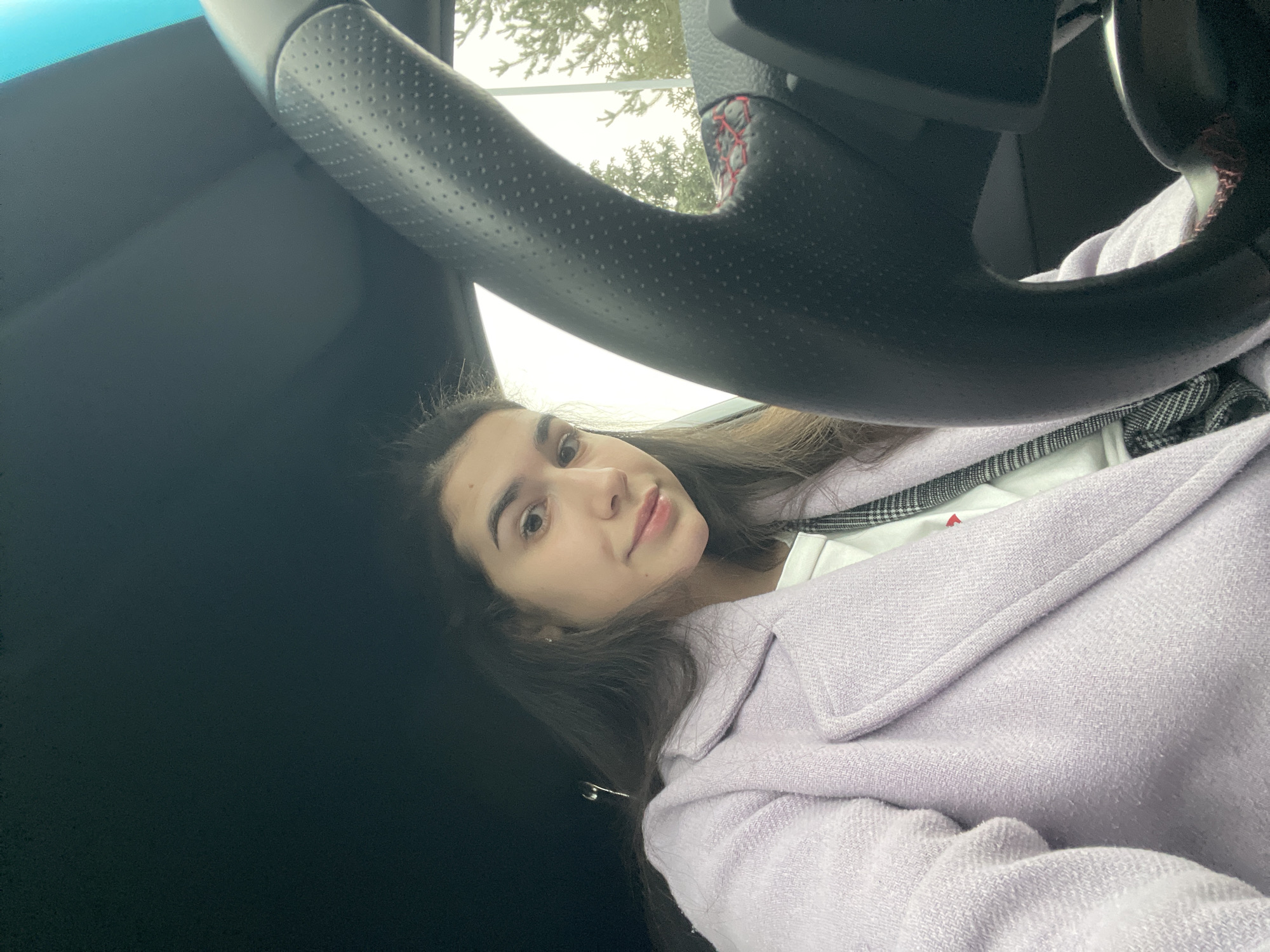 A woman is seated inside a car. The steering wheel of the vehicle is visible. The woman has brown hair and is wearing a lavender jacket over a white top with a plaid pattern. The car's interior is dark. The location appears to be outdoors during daylight hours. The city and country cannot be determined from the image.