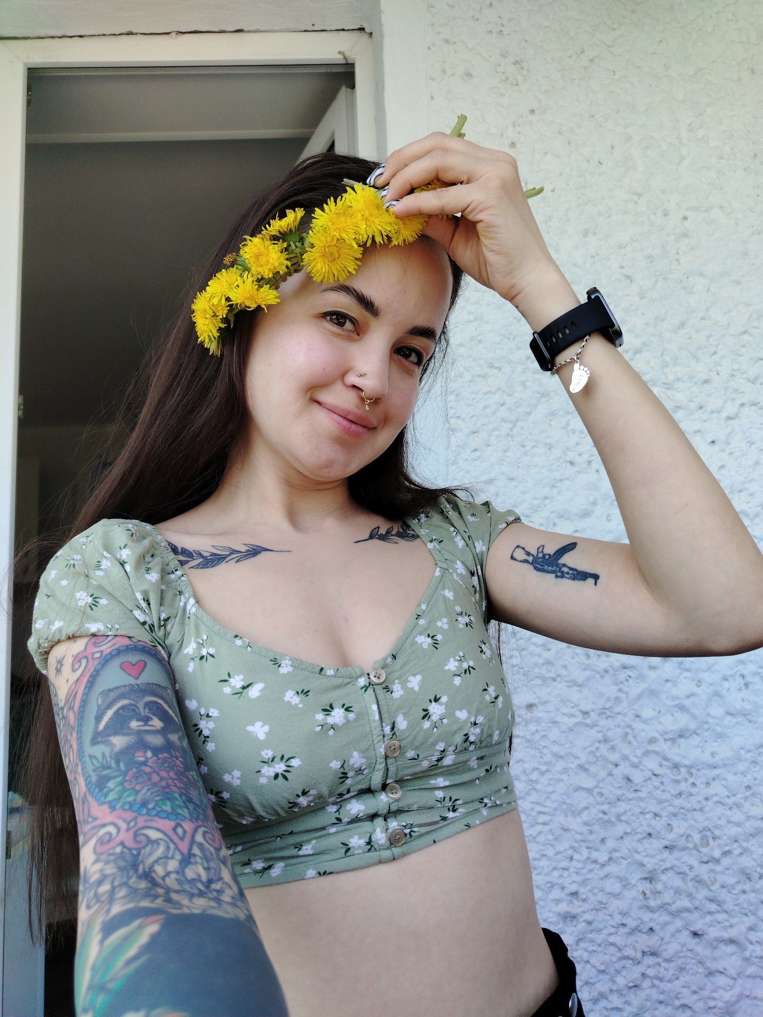 A woman is posing for a selfie outdoors. She has long, dark hair and is wearing a floral crop top. A crown of yellow dandelions adorns her head, and she is holding another dandelion. She has multiple tattoos on her arms and chest, and piercings in her nose. She is wearing a smartwatch with a charm. The background includes a white door and a textured white wall. The location details are not discernible in this image, therefore, I can't identify the location as Noviy, Russia.