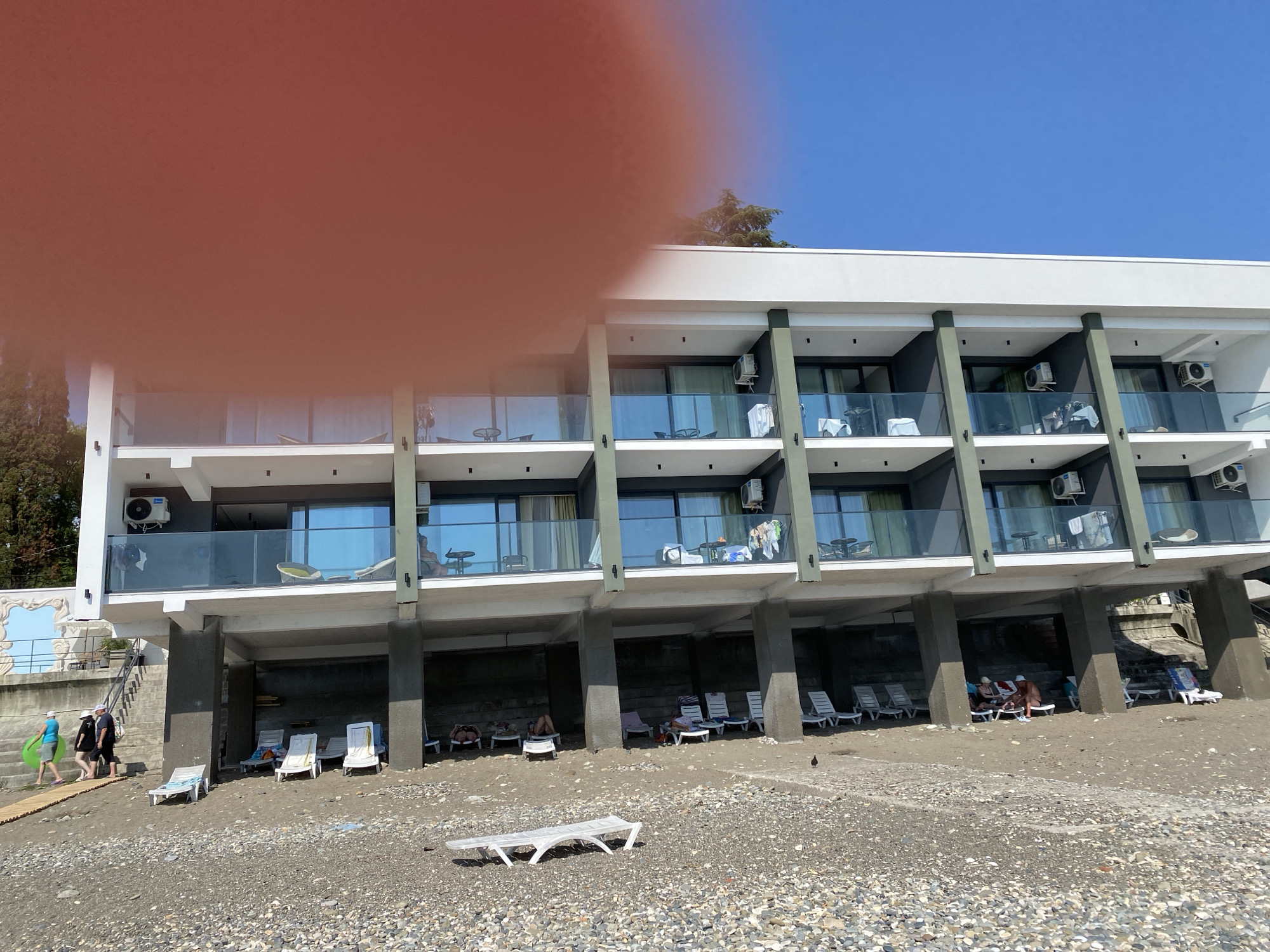 The image shows a multi-story hotel building on a pebble beach in Chabkwara, Abkhazia. The hotel has balconies with glass railings and air conditioning units visible on some balconies. Below the hotel, there is a covered area with concrete pillars, where several lounge chairs are placed. On the beach, there are more lounge chairs, some occupied by people. In the foreground, there is a white plastic lounge chair. A group of people is walking on a wooden pathway towards the hotel. The sky is clear and blue.