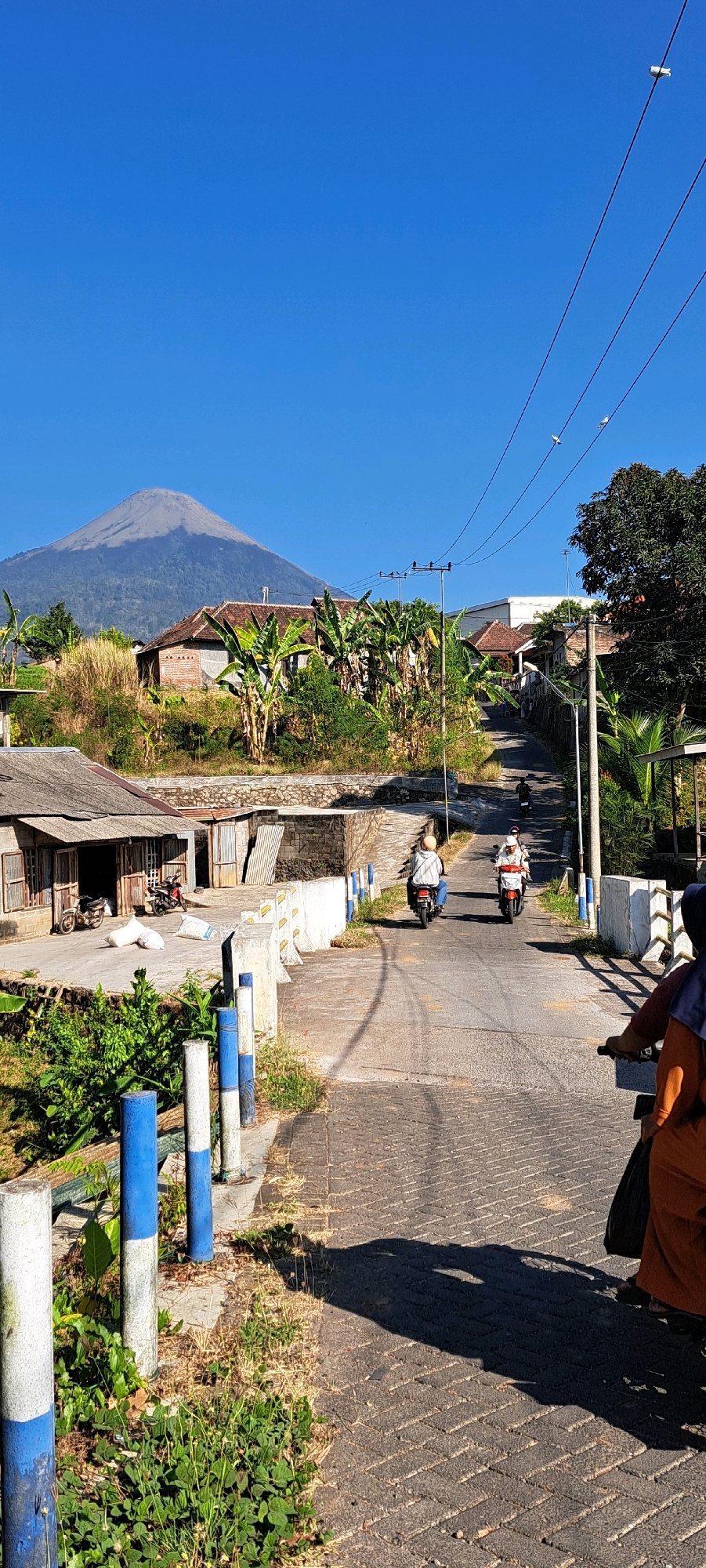 The image depicts a view of a road in the city of Sumber Suko, Indonesia, leading towards a mountain. On the left side of the road, there is a low-lying building with a corrugated metal roof and a wooden facade. Adjacent to this building are stacks of bagged materials and two motorcycles. Further up the road, there is a bridge with white and blue striped bollards along its side. Several individuals are seen riding motorcycles on the road, moving away from the viewer. The road is lined with trees and vegetation, including banana trees. In the background, houses with tile roofs are visible, nestled among lush greenery. A large, majestic mountain dominates the horizon under a clear blue sky. Electrical wires run across the top of the frame. The scene appears to be an everyday street view with local transportation and residential structures.