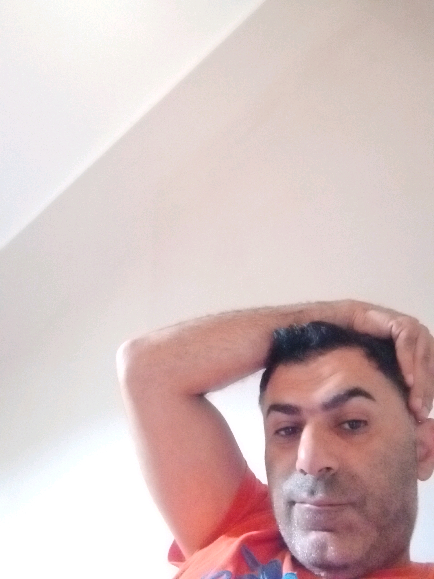 The image is a close-up, low-angle selfie of a man with dark hair and stubble. He is wearing an orange t-shirt. His right arm is raised and his hand is resting on the back of his head. The background appears to be a plain, light-colored wall with a diagonal shadow. There is no visible indication of the city of Al-Obour, Egypt, in the image.