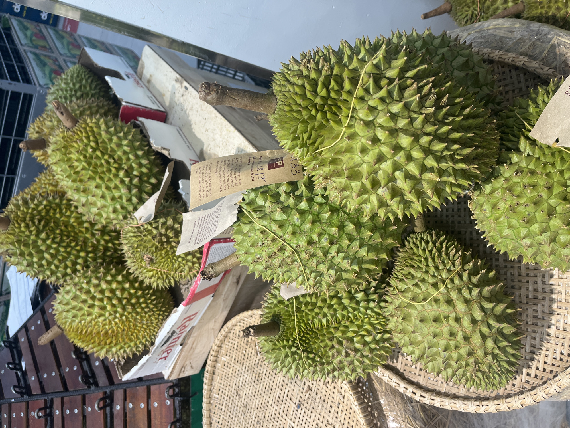This is a medium shot, eye-level, outdoor photograph of several durians. The durians are large, green, and spiky, with some having yellow strings tied around them. Some durians are in a woven basket, while others are stacked on top of each other or placed on a metal grate structure. In the background, there are cardboard boxes and what appears to be a white wall. Some text is visible on a paper tag attached to one of the durians, which seems to be a label or information about the fruit. The lighting suggests it is daytime, likely sunny, given the shadows cast. The scene appears to be a market or roadside stall selling durians in Nha Trang, Vietnam. There are no people visible in the frame, and the focus is entirely on the fruit.