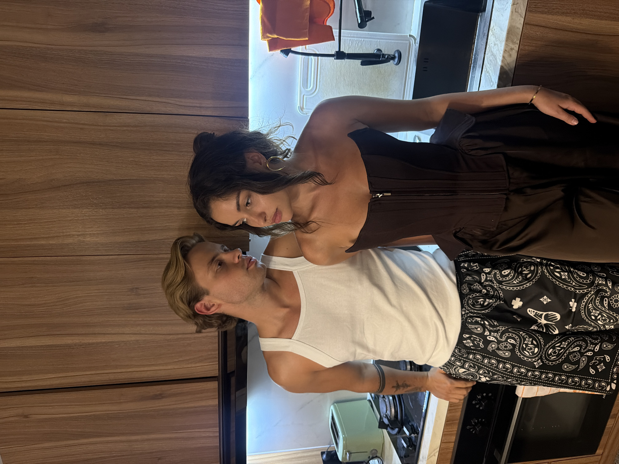 A woman and a man are standing close to each other in a kitchen. The woman has dark hair pulled into a bun and is wearing a brown corset top. The man has blond hair and is wearing a white tank top and black bandana-patterned shorts. They are both looking at each other.
The kitchen has wooden cabinets, a white countertop, and a stainless steel sink. There is a toaster oven on the counter next to the man.
The location is Kerobokan, Indonesia.