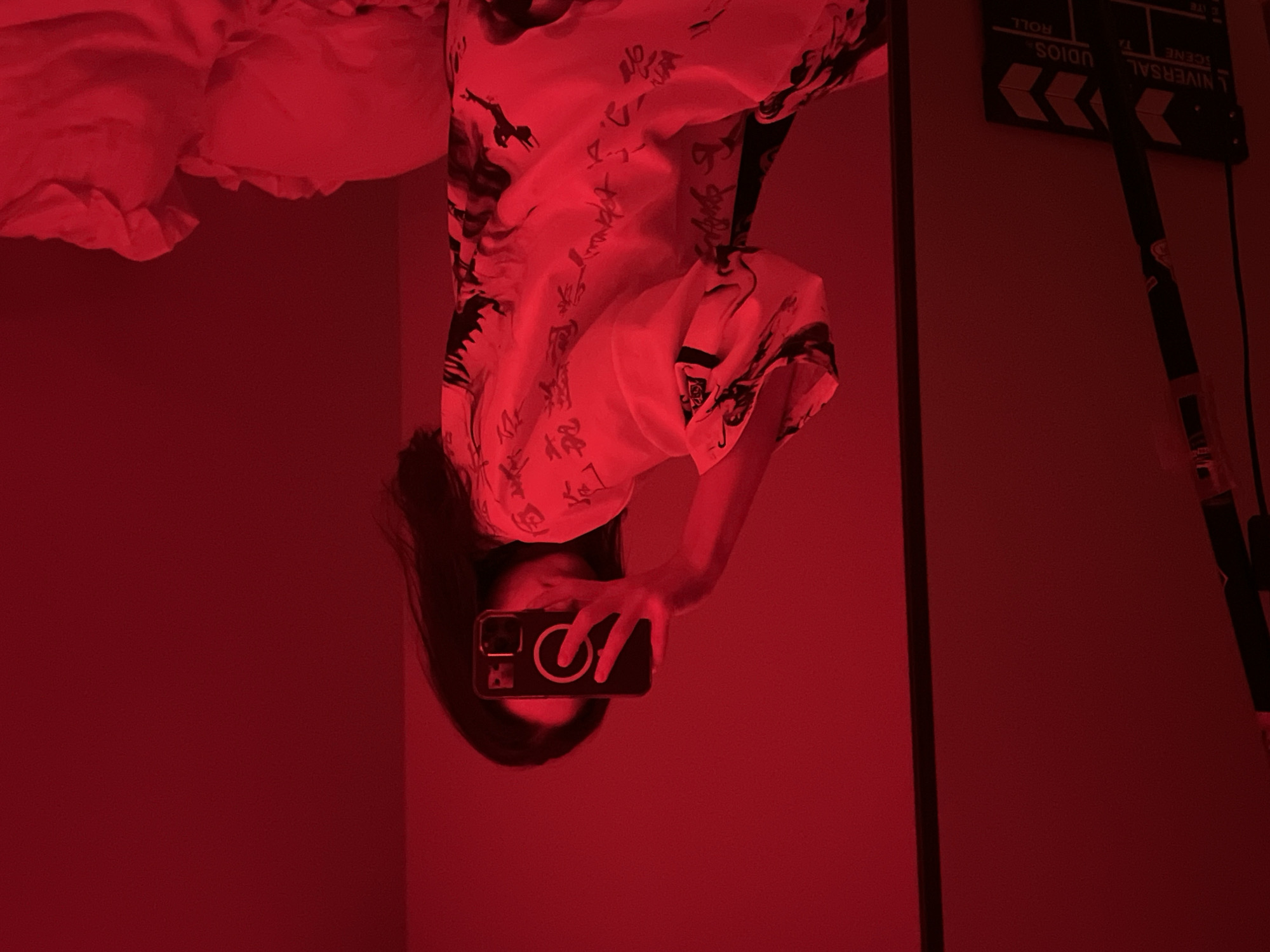A person, seen in a mirror reflection, is taking a selfie with a smartphone held horizontally. The person is wearing a white robe with black oriental-style prints. The scene is lit with a strong red light, casting a red hue over everything. To the left, a film clapperboard with "UNIVERSAL STUDIOS" printed on it is visible. The background appears to be a bedroom, with pillows visible on the right. The image does not contain any identifiable landmarks or context that would place it in Ternopil, Ukraine.