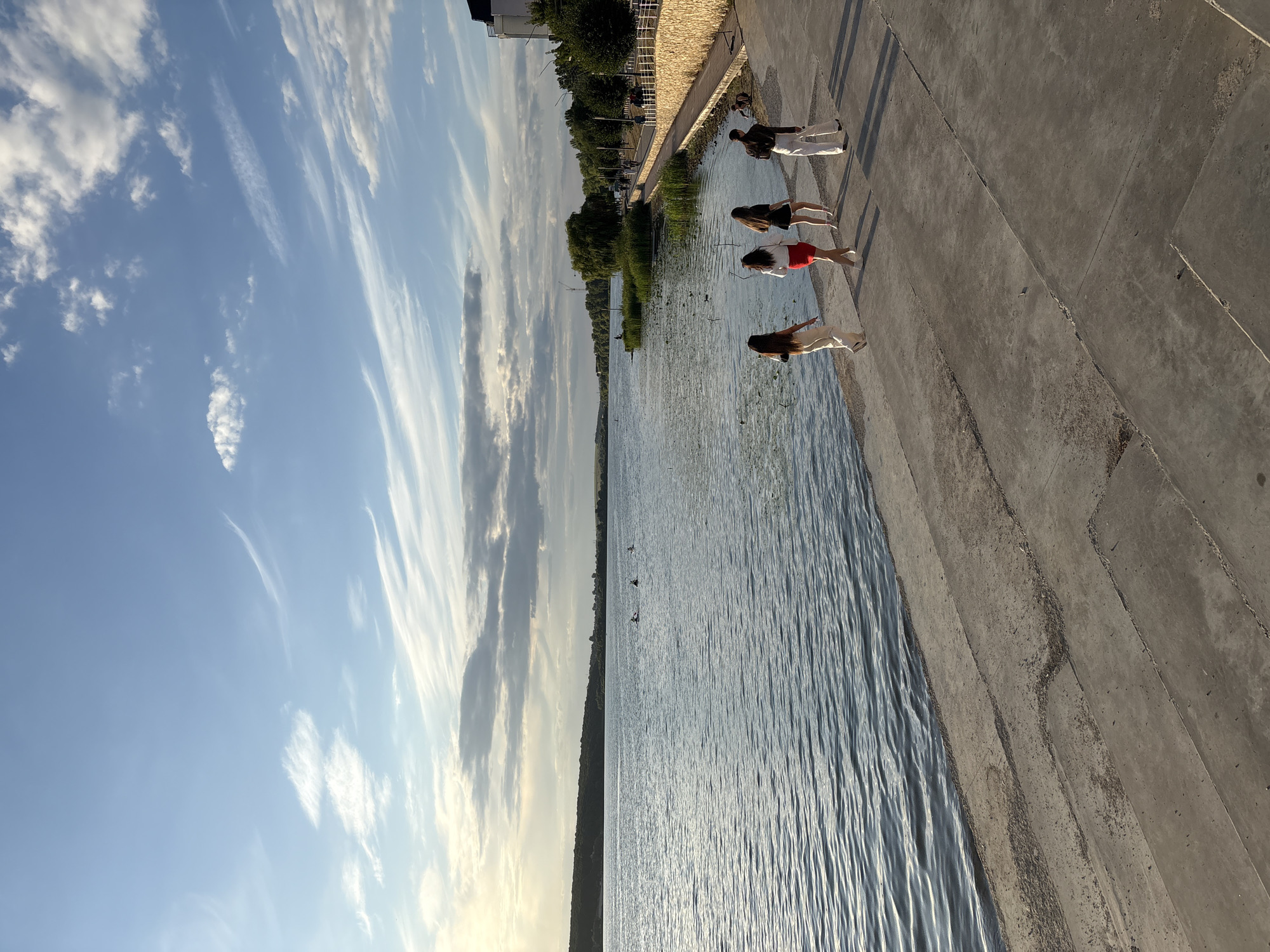 The image captures a scene along the Ternopil Lake promenade in Ternopil, Ukraine, during what appears to be the late afternoon or early evening. The sky is a clear blue with scattered wisps of white clouds, and the sun is low on the horizon, casting long shadows.
In the foreground, a group of four young women are walking along the concrete embankment. The woman closest to the viewer is wearing cream-colored pants and a white top. To her right, another woman is in a red skirt and white top. A third woman wears a black dress, and the fourth is dressed in white pants and a dark top. Their movement is directed away from the viewer and towards the right side of the frame.
The Ternopil Lake stretches across the middle ground, its surface rippling gently. In the distance, a tree-lined shore forms the horizon, with a few buildings visible to the far right. Several small boats or kayaks are dotted on the water further out, and a few ducks are swimming closer to the shore.
The promenade itself is paved with stone and concrete, with a low wall separating it from the lake. The overall impression is one of a pleasant, leisurely stroll in a scenic urban setting.