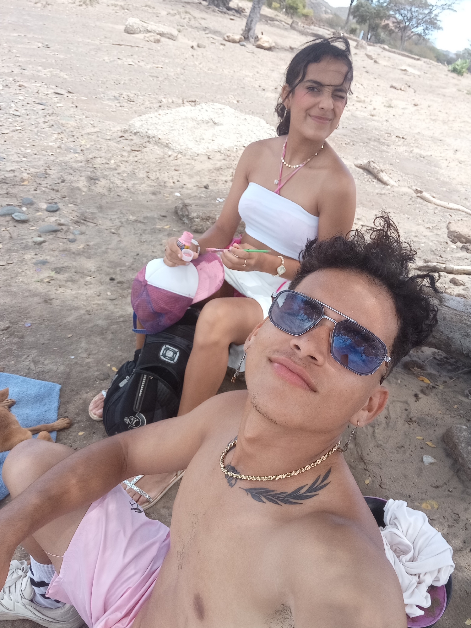 The image shows a man and a woman posing for a photo on a beach. The man is in the foreground, shirtless and wearing pink shorts, sunglasses, a gold chain, and sneakers. He has a tattoo on his left collarbone. The woman is seated behind him, wearing a white tube top and skirt. She is winking at the camera and holding a pink and white baseball cap. A black backpack, a blue towel with a dog, and some clothing are visible near them. The background shows a sandy beach with sparse vegetation and a few trees. The location is likely in Altagracia, Venezuela.