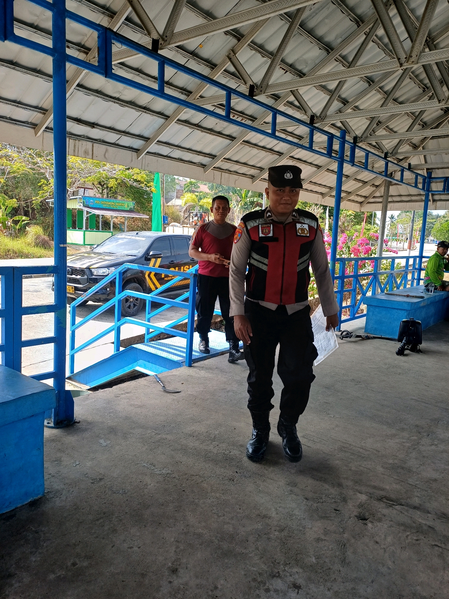 In the city of Pasir Belengkong, Indonesia, a policeman in uniform walks towards the camera, holding a document. He is wearing a black cap, a red and black vest over a gray shirt, and black pants with black boots. Behind him, another person in a red shirt and black pants stands near a black police vehicle with yellow and black markings that read "POLISI" and "POLSEK PASER". The vehicle is parked in front of a small green and white building with signage. The scene is outdoors, under a covered structure with a metal roof supported by blue metal poles. Blue railings and steps are visible, as is a concrete floor. In the background, lush greenery and trees are present, along with some pink flowers. Another person wearing a green shirt is seated on a blue bench to the right, with a table and a glass in front of them. A black backpack is on the ground near the seated person.