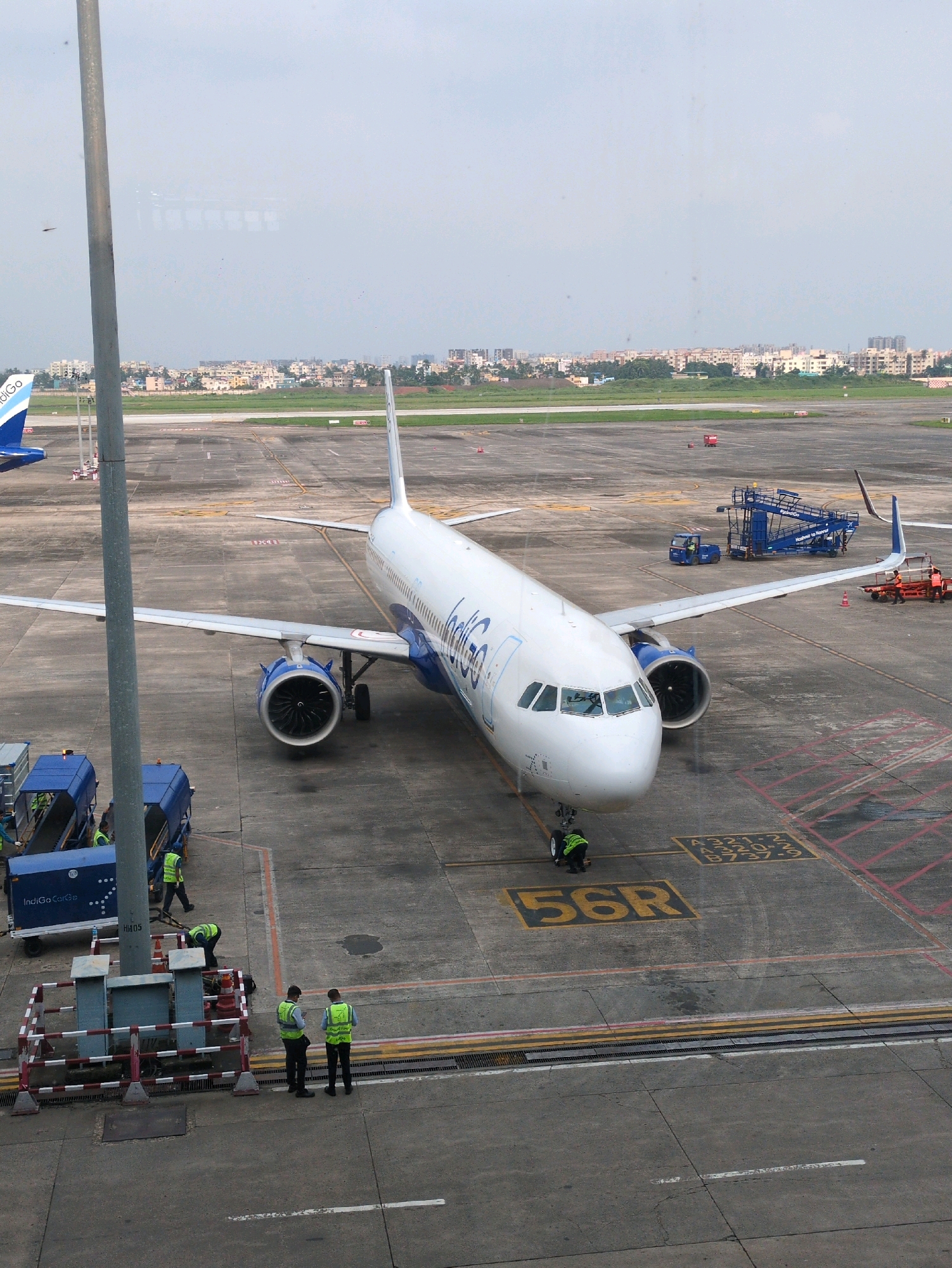 An Indigo Airlines airplane is parked on the tarmac at Netaji Subhas Chandra Bose International Airport in Dum Dum, India. The plane is white with blue accents and the Indigo logo is visible on its side. Several airport personnel in high-visibility vests are working around the aircraft, including one person near the front landing gear and others by a baggage conveyor belt. A mobile stairway is positioned next to the plane, and other ground support equipment is visible in the background. The airport tarmac is marked with yellow lines and numbers, indicating parking positions. In the distance, buildings and trees form the cityscape of Dum Dum. The sky is overcast.