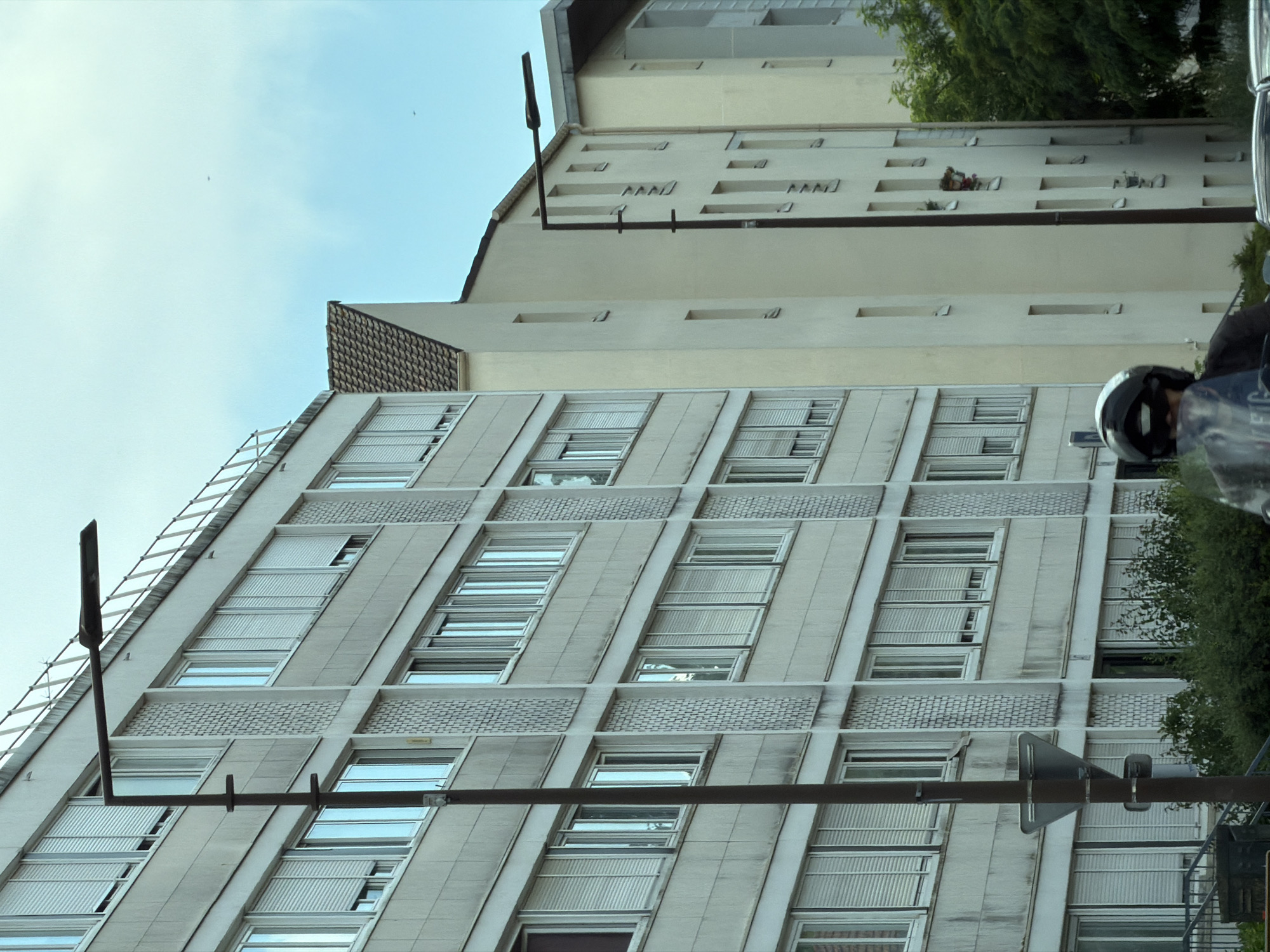 The image depicts an urban scene in Vélizy-Villacoublay, France, focusing on the architecture of apartment buildings and a street. In the foreground, a person on a motorcycle, wearing a helmet, is partially visible. The motorcycle's windshield is in the lower part of the frame.
Dominating the left side of the image is a multi-story apartment building with a grey, textured facade. It features rows of rectangular windows, many of which are covered by white slatted blinds. The building has a modern, utilitarian design. A dark-colored lamppost with a horizontal light fixture extends across the frame in front of this building, obscuring some of the windows. A triangular traffic sign is attached to the lamppost.
To the right of the grey building, a lighter-colored, taller apartment building is visible, with a cream-colored facade. This building also has rows of windows, some of which appear smaller and more spaced out than those on the grey building. A pitched roof with brown tiles is visible at the top of this building, suggesting it might be an older section or a different architectural style. Further to the right, another building with a grey upper section and lighter walls is seen, partially obscured by trees.
The sky is overcast with a pale blue hue, indicating a cloudy day. Green foliage from trees is visible at the bottom and right edges of the image, suggesting a residential area with some greenery. The overall impression is that of a typical urban residential environment.
