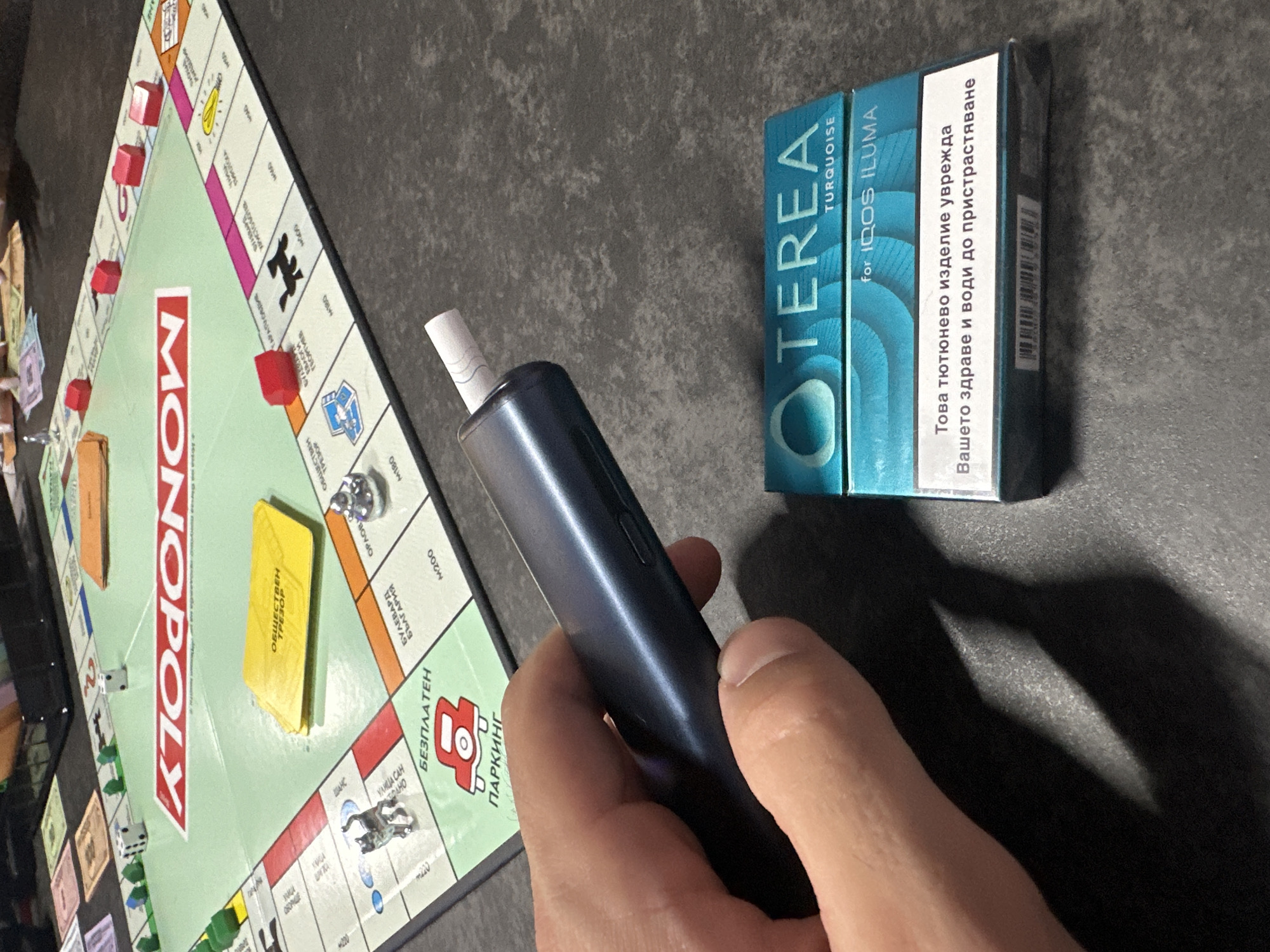 The image shows a close-up of a person's hand holding a dark blue electronic smoking device. In the background, a Monopoly board game is visible. The board is set up with various properties, tokens, and dice. A pack of TEREA Turquoise for IQOS ILUMA is also present in the foreground, next to the Monopoly board. The environment appears to be an indoor setting with a dark, textured surface that serves as the backdrop. The Monopoly board is mostly green and cream colored, with red and yellow accents. The electronic device is cylindrical and metallic. The TEREA pack is rectangular and predominantly teal colored with white text. The overall scene suggests a casual moment of relaxation or play.
