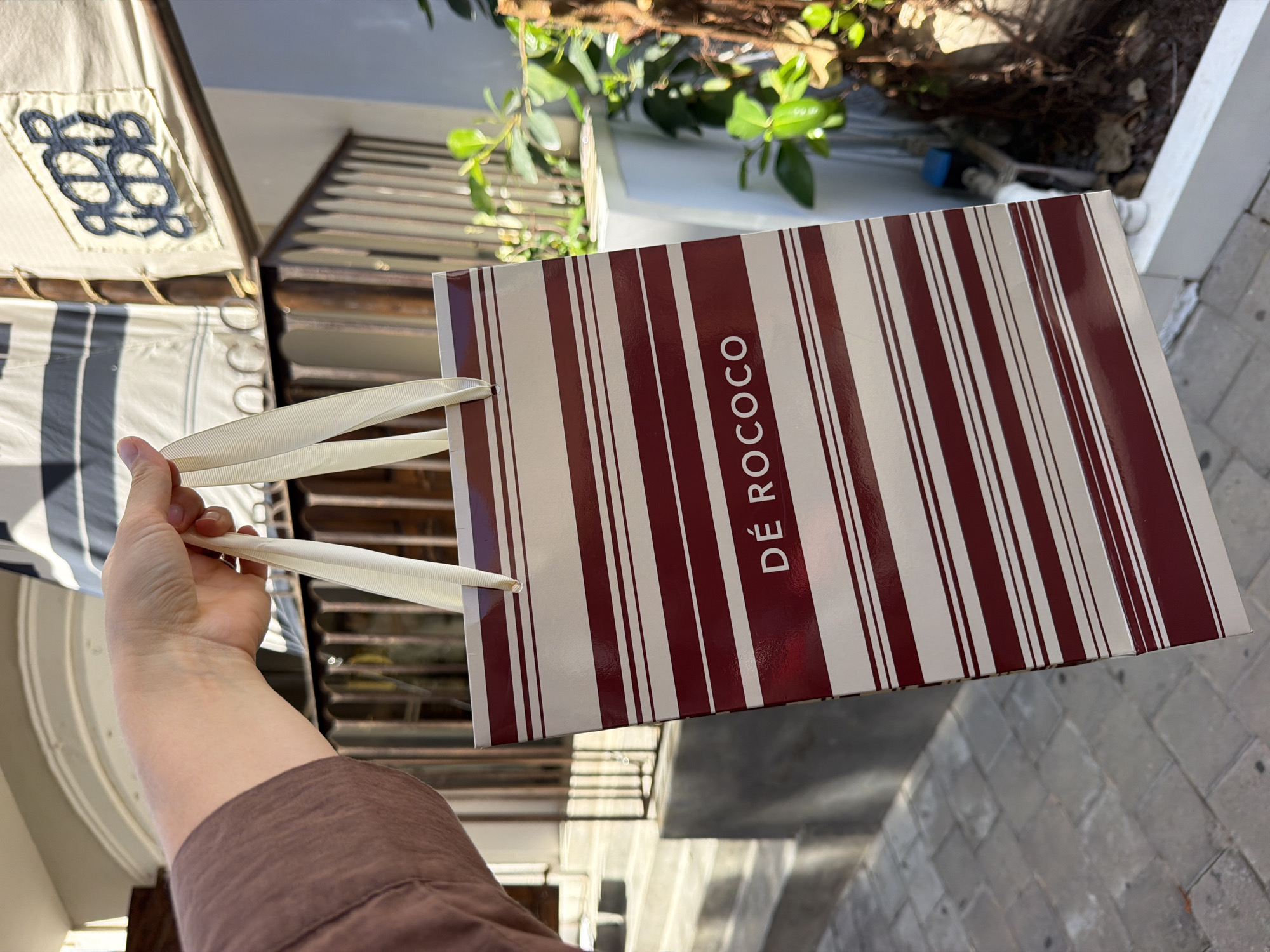 A person's hand, wearing a brown shirt, holds a shopping bag with a striped pattern of maroon and white. The bag displays the text "DE ROCOCO" in white lettering. In the background, a building with a railing and plants is visible. The ground is paved with rectangular stones. The scene appears to be outdoors in an urban setting.