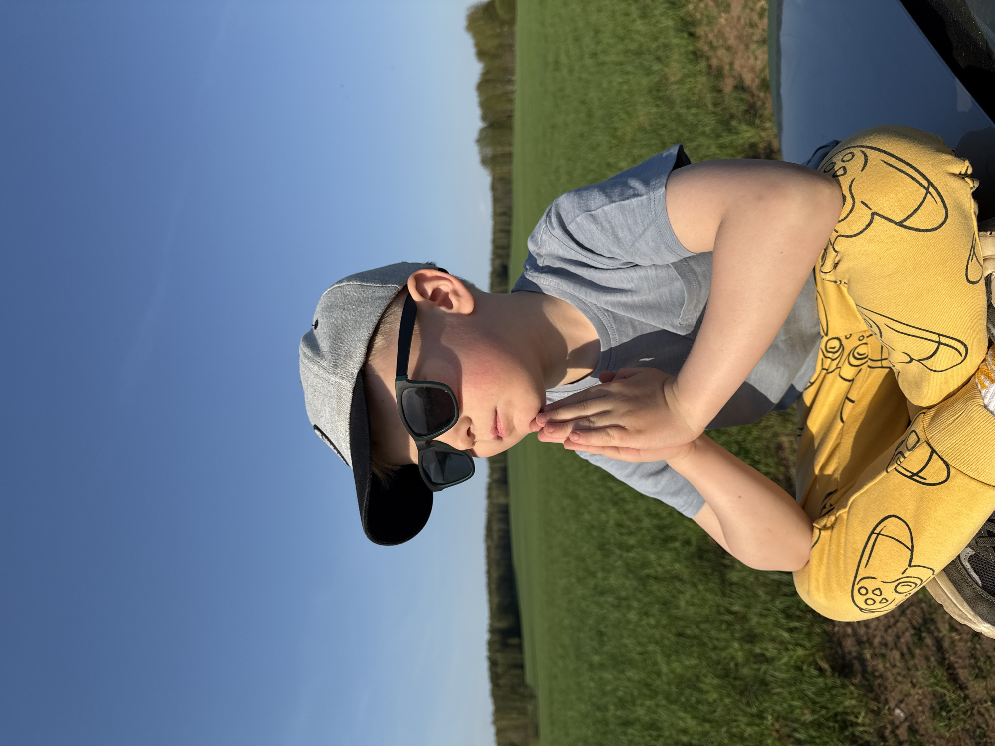 A child wearing a grey baseball cap, sunglasses, and a light blue t-shirt is crouching on a dark-colored car, with his hands clasped together. He is also wearing yellow pants with a controller print on them. The background consists of a green field, a line of trees, and a clear blue sky. The location is Шумиха, Russia.