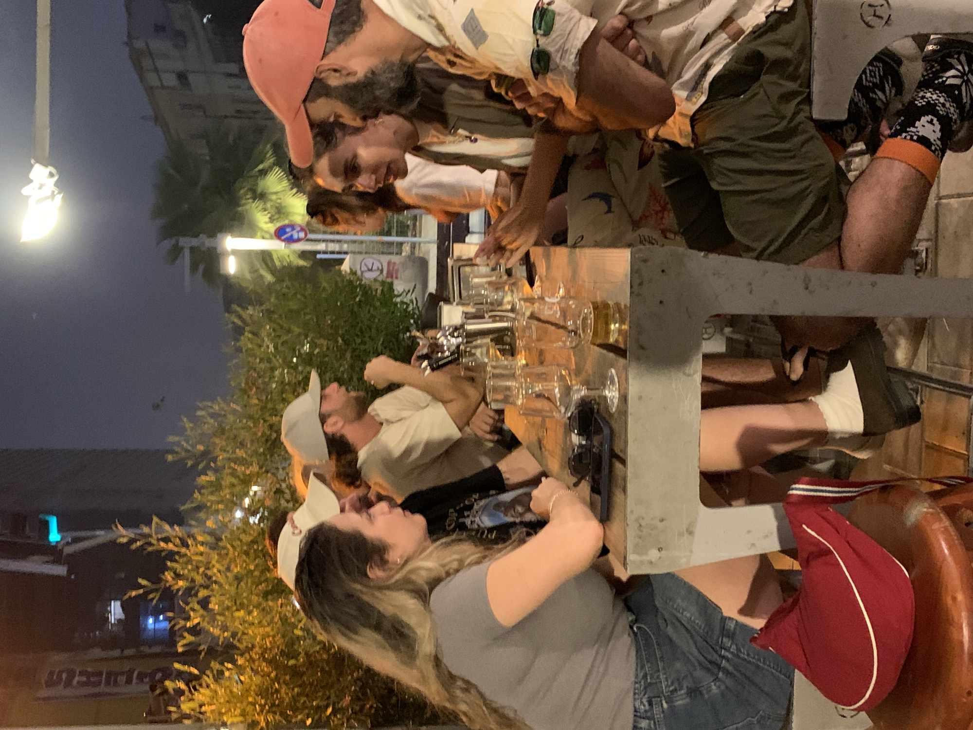 A medium shot depicts a group of people seated at a long communal table outdoors, possibly at a restaurant or bar. The setting appears to be in Tel Aviv, Israel, based on architectural styles. The sky is dark.
From left to right:
The woman closest to the foreground is wearing a light gray shirt, a denim skirt, a white baseball cap, and has long, blonde highlighted hair. Her body is turned towards the table but her head is turned to the left.
The person beside her is wearing a white T-shirt and a gray baseball cap. The only other person clearly visible in the group is a man with a peach-colored baseball cap. He is wearing an patterned shirt and green shorts.
The table has numerous empty beer glasses along its length, implying recent or ongoing consumption.
A red bag is hanging on the chair that the woman is sitting on.
The setting seems to be a lively urban environment. Buildings and streetlights are visible in the background.