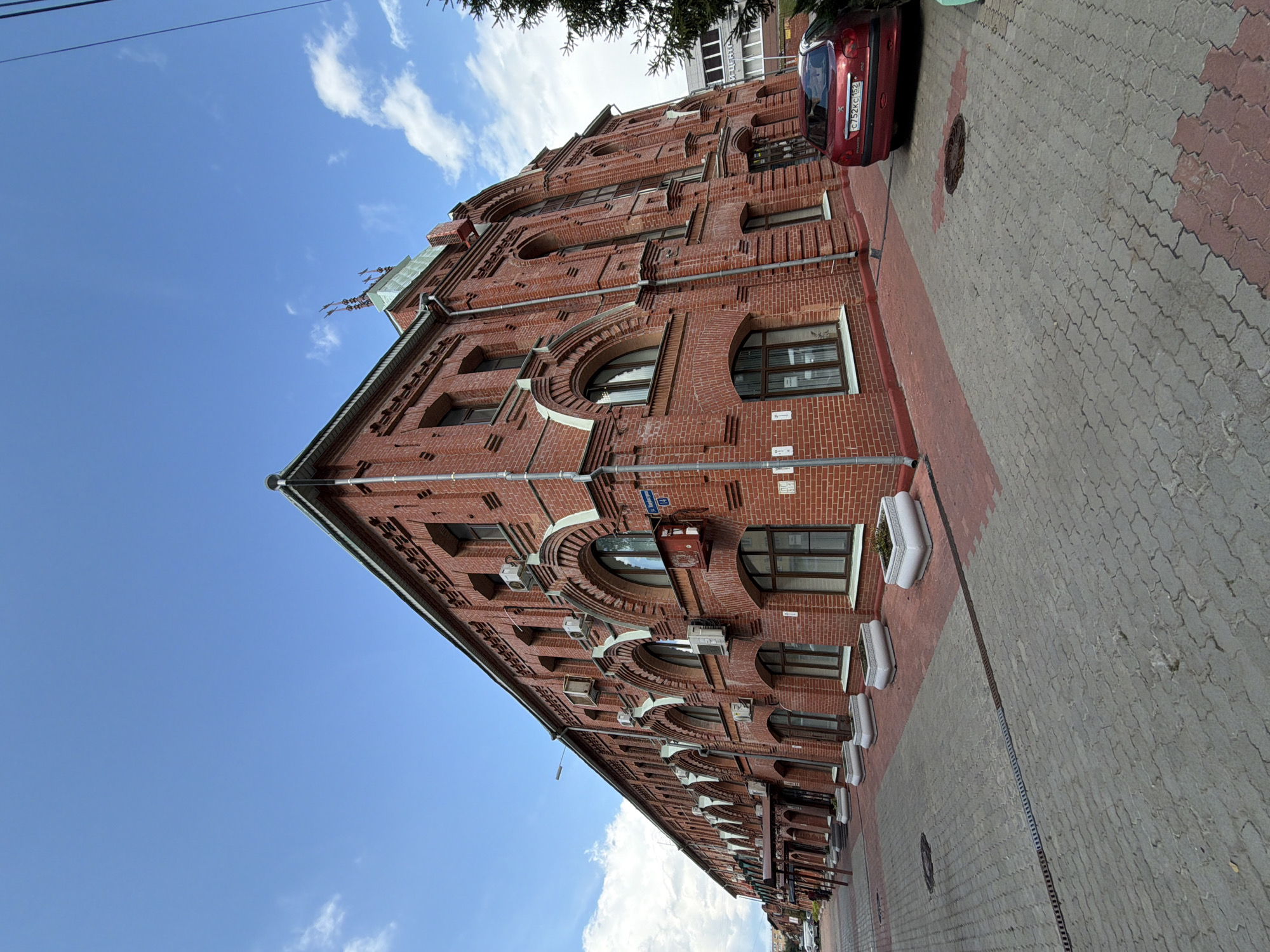 The image shows a red brick building on a sunny day. The building has several floors with arched windows. There are air conditioning units mounted on the exterior walls. The street in front of the building is paved with grey and red interlocking pavers. A red car is parked on the right side of the street. The sky is blue with some white clouds. In the background, there are some trees and other buildings. The street sign visible on the building reads "Ул. Добролюбова 14", which translates to "Dobrolyubov St. 14". This indicates the location is Dobrolyubov Street, number 14, in Nizhny Novgorod, Russia. There are no people visible in the image.