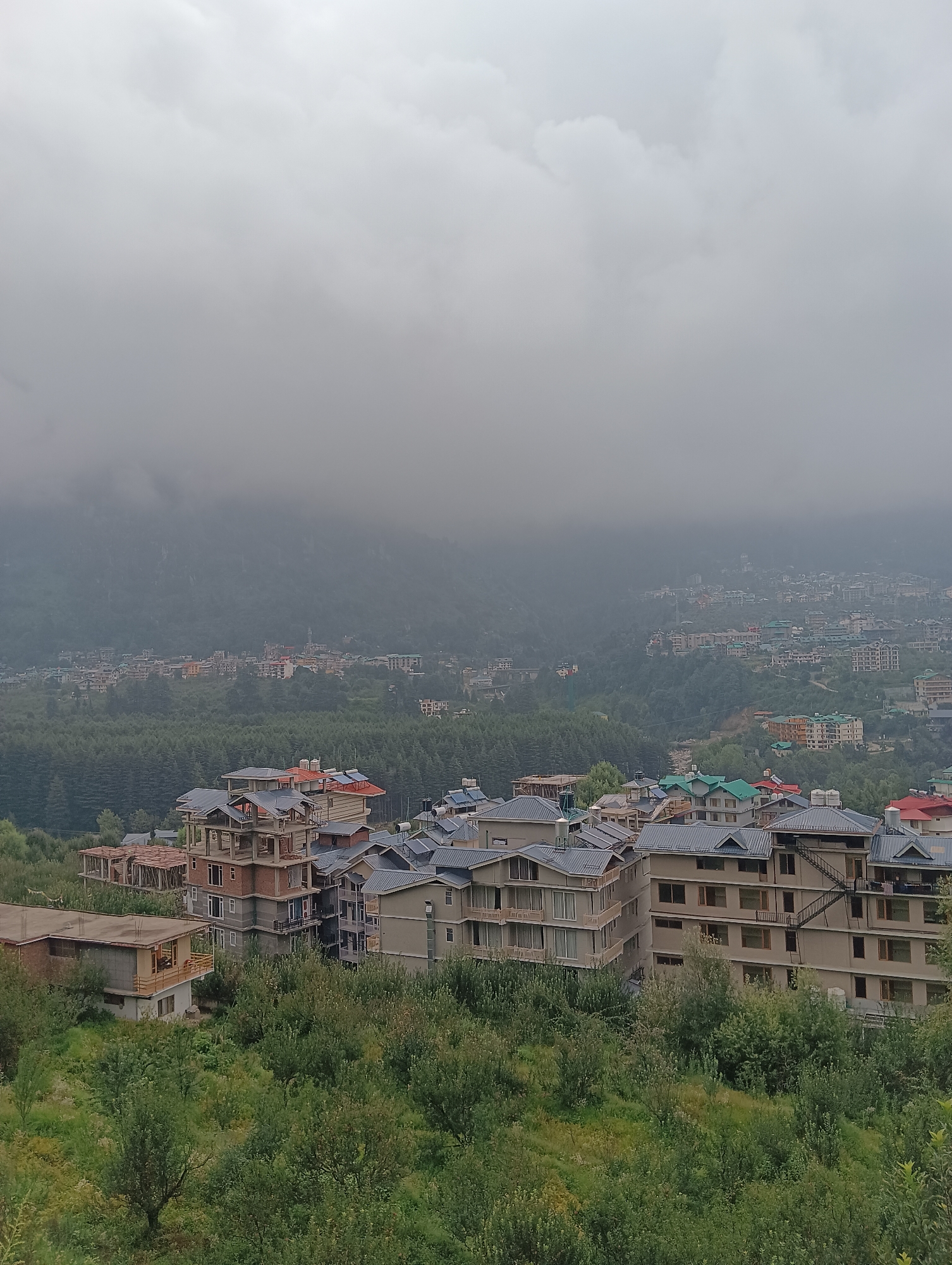 The image depicts a hazy, overcast day in Siumsa, India. In the foreground, several multi-story buildings with peaked roofs are visible, some appearing under construction. These buildings are nestled amongst lush green trees and vegetation. In the middle ground, a dense forest of pine trees stretches across the landscape, and further beyond, a town or city is spread out across rolling hills. Numerous buildings with various colored roofs are visible, and the distant mountains are shrouded in thick fog or clouds. The overall impression is of a scenic, mountainous region experiencing inclement weather.