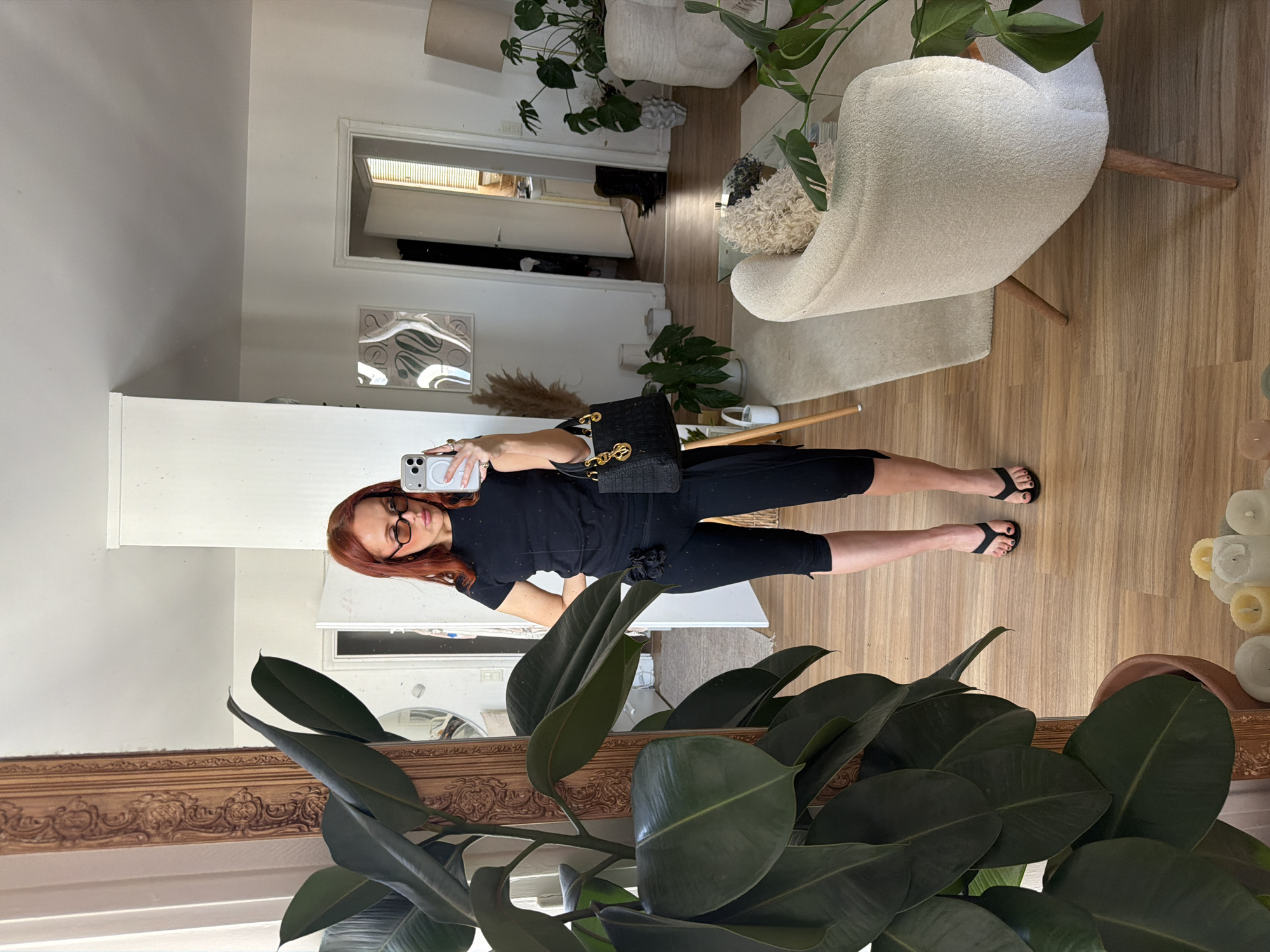 A woman with red hair and glasses takes a full-body selfie in a mirror. She is wearing a black top and black shorts, and black sandals. She is holding a smartphone in her right hand and a black handbag hangs from her left shoulder. The reflection reveals a room with wooden flooring and white walls. There's a white chair with wooden legs, several potted plants, a doorway leading to another room, and various decorative items. The mirror itself has an ornate wooden frame. The location cannot be identified as being in Paris, France.