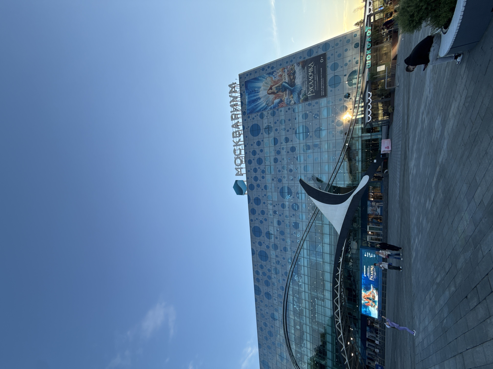 The image depicts the Moskvarium (Moscow Oceanarium) building in Moscow, Russia, under a clear, sunny sky during the late afternoon or early evening. The prominent building features a modern glass facade patterned with various blue circles, reflecting an aquatic theme. A large vertical sign on the building clearly spells out "МОСКВАРИУМ" in Cyrillic.
A substantial billboard on the upper part of the building showcases an illustration of a mermaid and advertises "РУСАЛОЧКА" (Rusolochka - The Little Mermaid) as a "МЮЗИКЛ НА ВОДЕ" (musical on water) "С МОРСКИМИ ЖИВОТНЫМИ" (with marine animals), titled "ЛЮБОВЬ ДВУХ МИРОВ" (Love of two worlds), suitable for ages "0+". Another screen below features additional branding for "РУСАЛОЧКА." Near the entrance, a kiosk displays "вход" (entrance) and "TOP." Partial text "Шоколо" (likely from "Шоколадница" cafe) is also visible.
The scene includes a paved plaza in front of the building. Several people are present: one individual in a black jacket and light pants leans on a grey planter filled with greenery on the right. A small group of three to four people stands near the building's entrance, while another person in purple pants walks away from the main entrance area. An abstract, black and white architectural element or sculpture is situated near the building's base. The low sun creates a warm glow on the building's upper right side, and the sky is a vibrant blue with minimal cloud cover.
