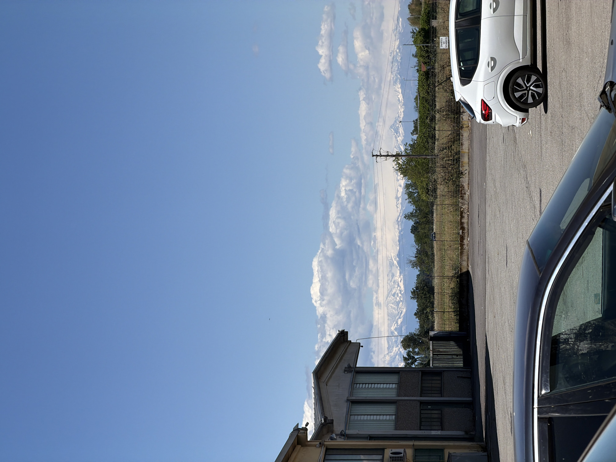 The image is taken in Gudo Visconti, Italy. A two-story building is in the foreground. Beyond the building, there is a view of mountains with snow-capped peaks. Clouds fill the space above the mountains. A fence and some trees are situated between the building and the mountains. A white car is parked on the right side of the image, and a black car is also partially visible in the foreground on the right. The sky is blue.