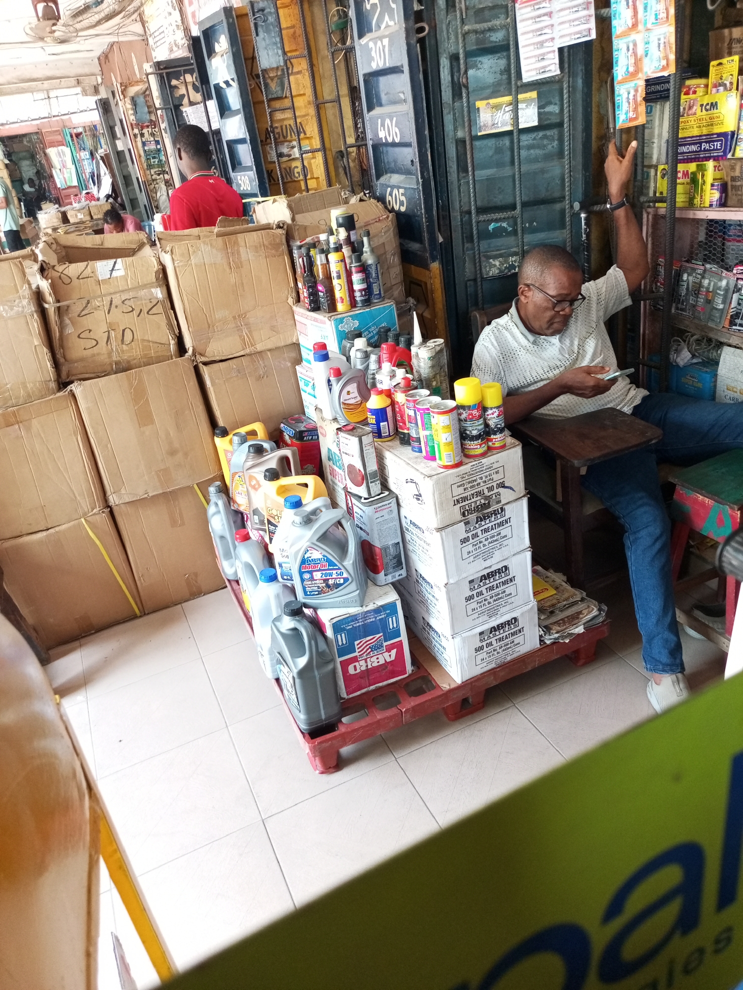 The image captures the interior of a bustling auto parts or general goods shop, likely a market stall, located in Satellite Town, Nigeria. The scene is brightly lit, indicating daytime.
In the foreground, a man wearing glasses and a light-colored, patterned polo shirt is seated on a wooden stool, engrossed in his mobile phone. His left arm is casually raised, resting on the top of a tall metal container that forms part of the shop's structure. His attention is focused downward on the device, suggesting a moment of leisure or waiting.
The shop is filled with a dense array of merchandise. To the left, numerous brown cardboard boxes are stacked high, some handwritten with labels such as "215,2" and "STD". In the center, a red plastic pallet holds an organized display of various automotive products, primarily large plastic containers of motor oil and oil treatments, including brands like "ABRO Motor Oil 20W-50" and "ABRO 500 OIL TREATMENT." Numerous spray cans and smaller bottles of lubricants and additives are also visible on top of cardboard boxes.
The background features robust metal containers serving as the shop's walls, with numbers like "307," "406," and "605" painted on them. One container also bears the word "LAGUNA". Shelves lining these walls are packed with other products, including "TCM EPOXY STEEL GUM" and "GRINDING PASTE". In the left background, another man in a red shirt is partially visible amidst further stacks of boxes, and a third person is faintly seen further in the distance. An old fan hangs from the ceiling. The overall impression is that of a functional, somewhat cluttered, yet organized commercial space.
