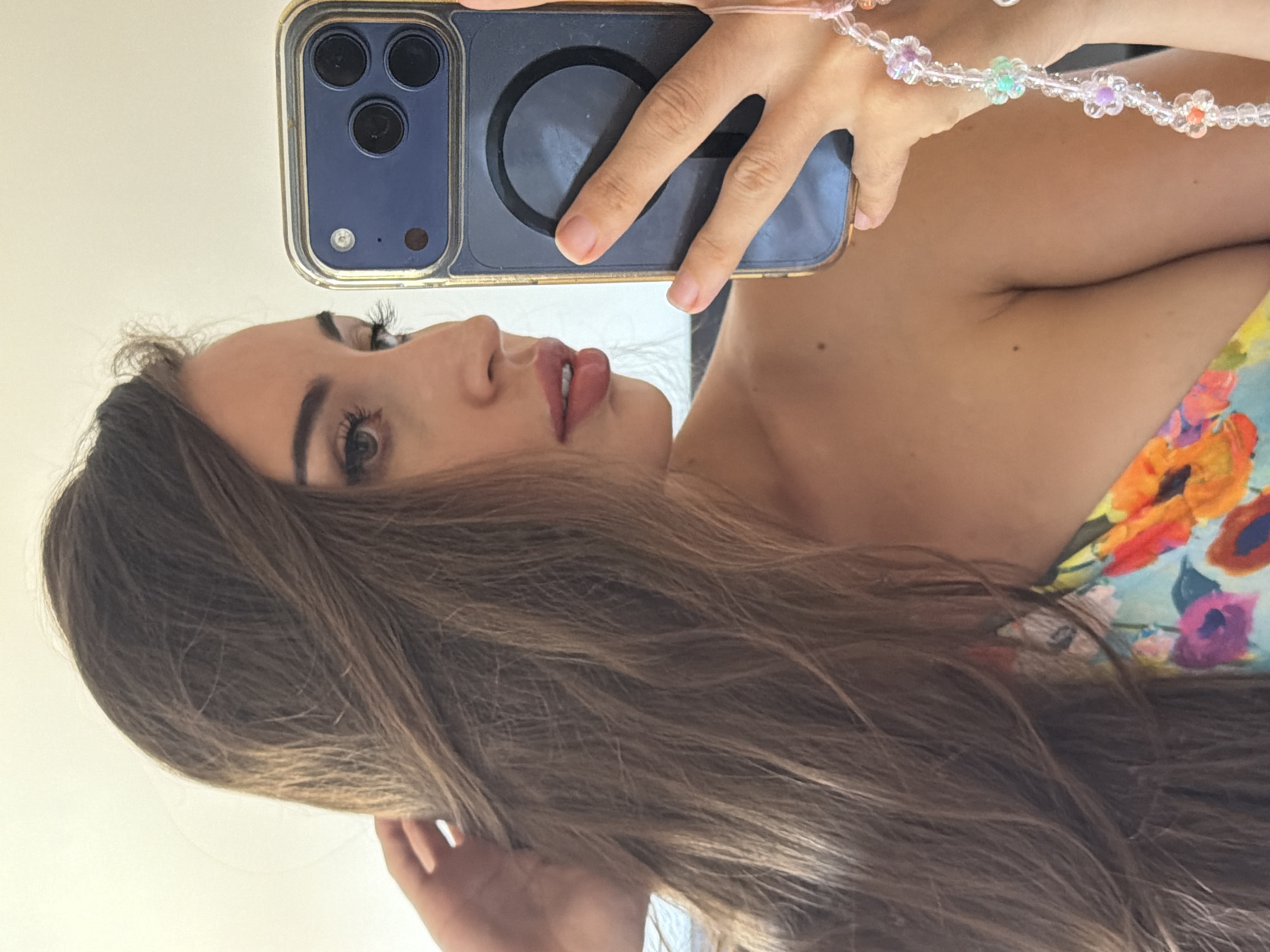 A young woman with long brown hair is taking a selfie in Tersefanou, Cyprus. She is wearing a colorful floral top and her left hand is holding a blue smartphone. A beaded charm bracelet is attached to her phone. The background is a plain, light-colored wall.